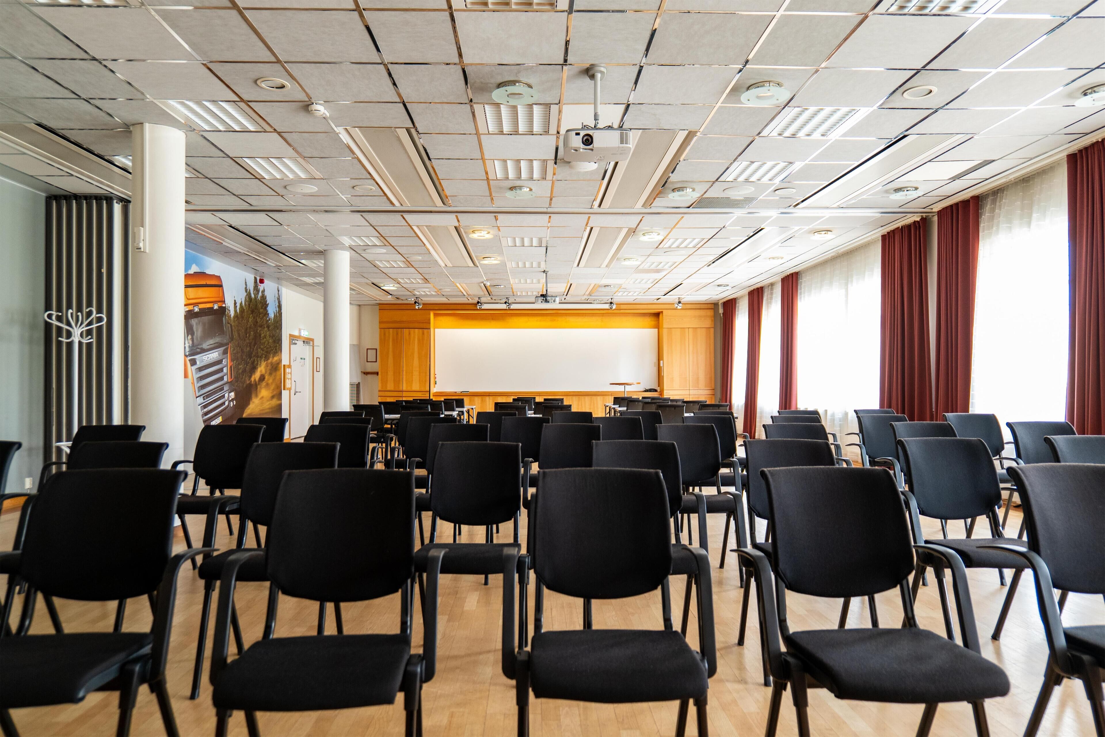 Konferenssihuone, jossa on riveissä mustia tuoleja, valkoinen projektorikangas ja puupanelilla verhoiltu seinä.