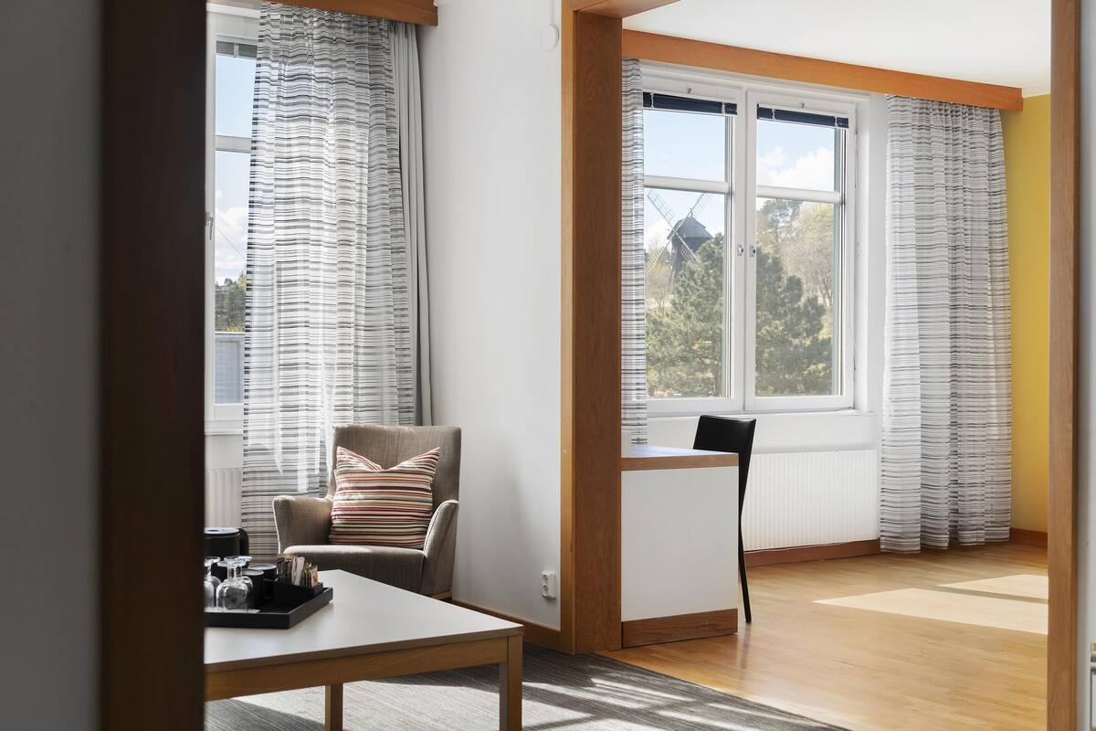 A room with a wooden-framed desk, a cushioned chair, a table with a tray, and large windows with curtains.