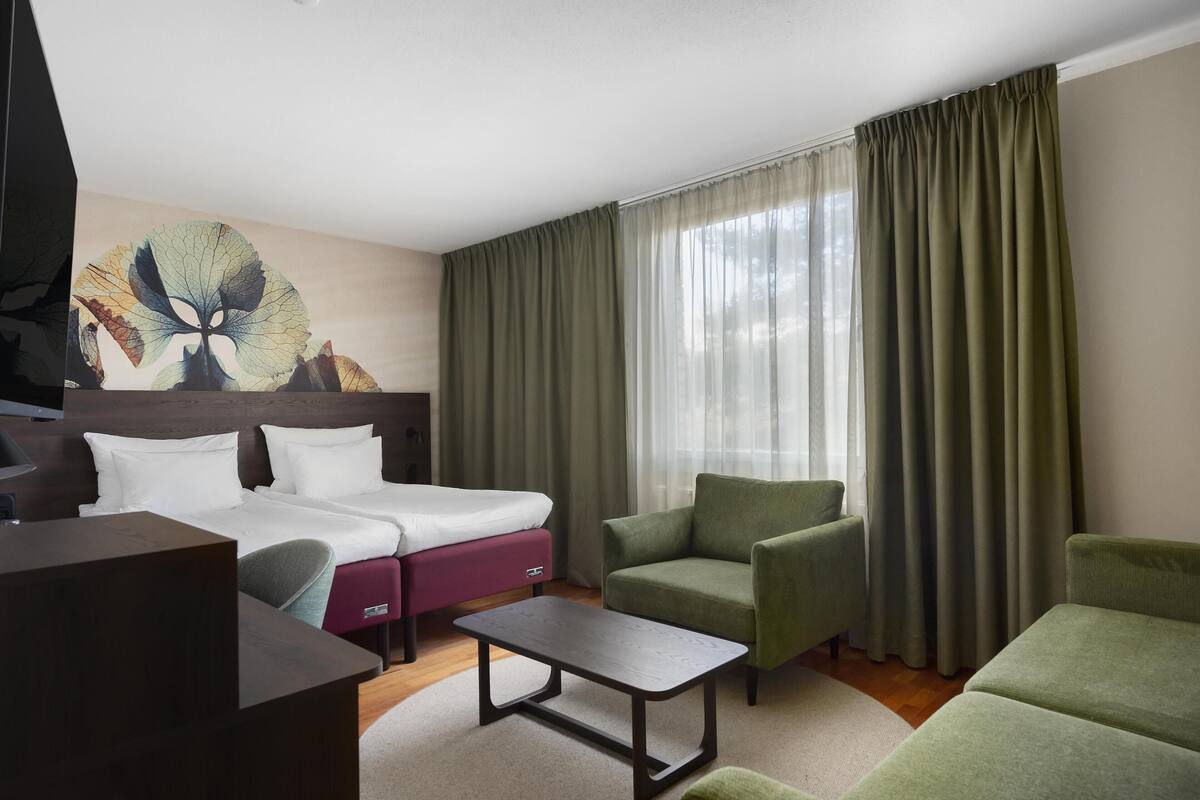 A hotel room with a bed, two green armchairs, a small table, and a painting on the wall.