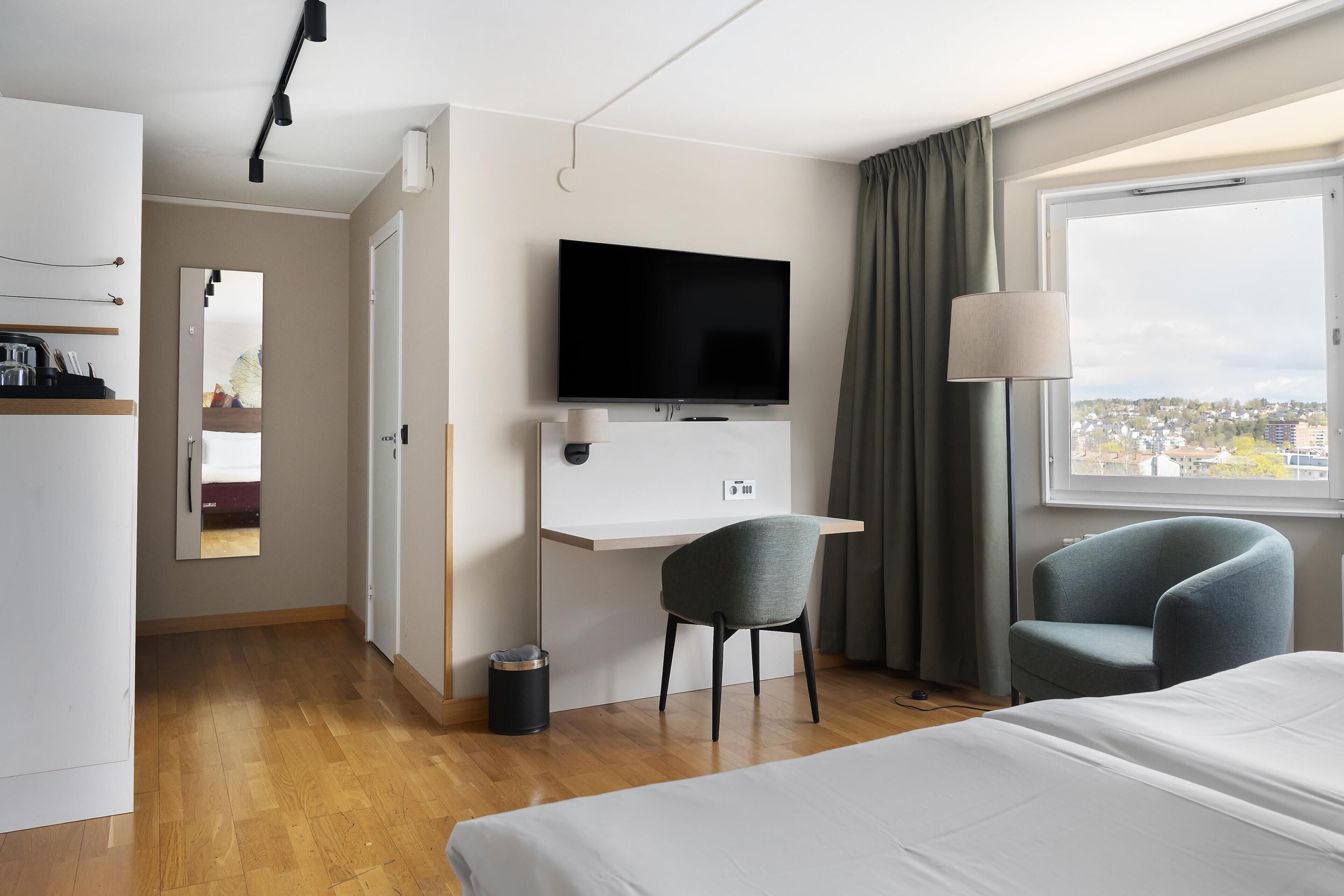 Et hotelværelse med en seng, et skrivebord med et fjernsyn, to stole, et vindue med udsigt og en dør til et andet værelse.