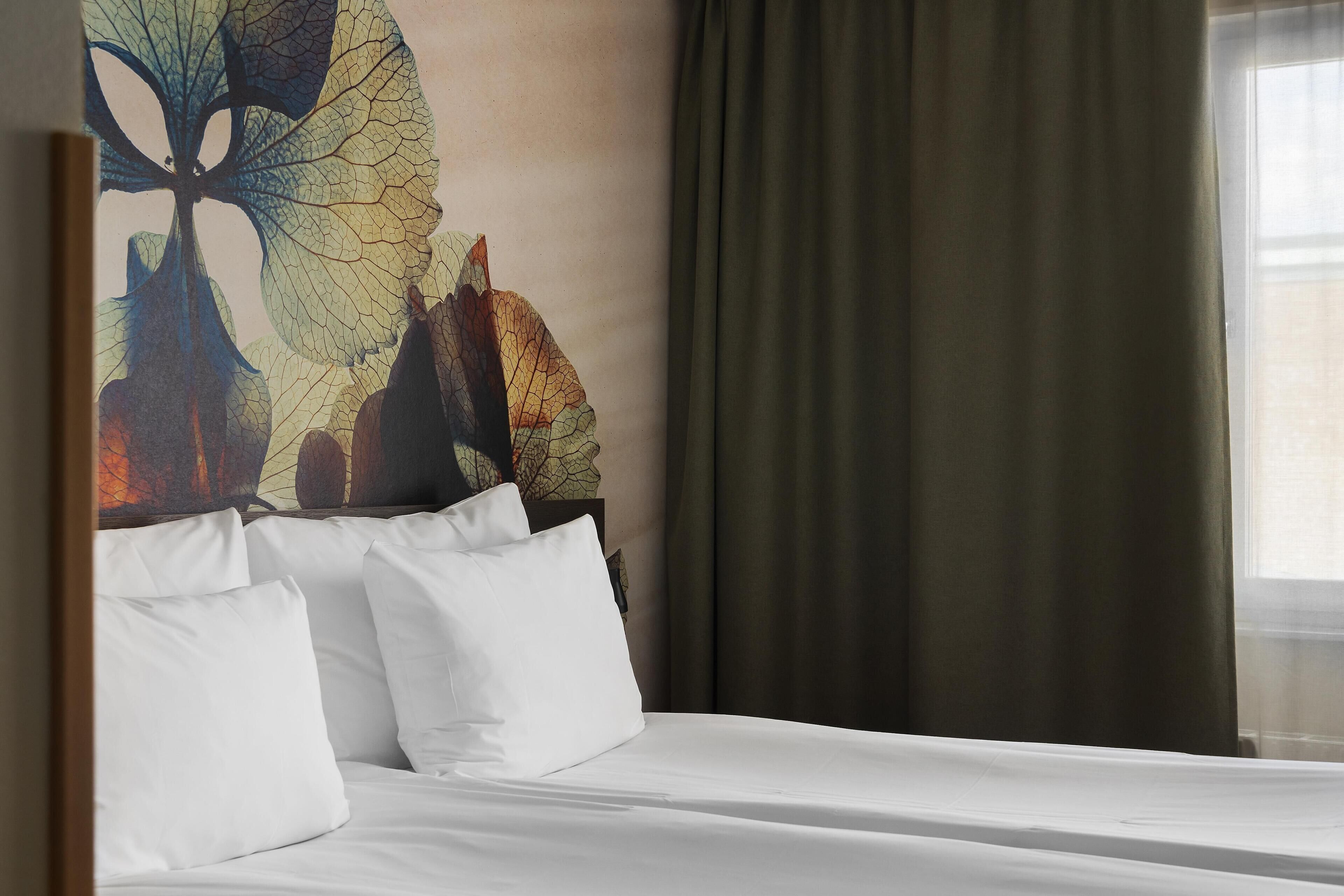 A hotel room with a bed, white pillows, a large leaf-patterned artwork on the wall, and a window with curtains.