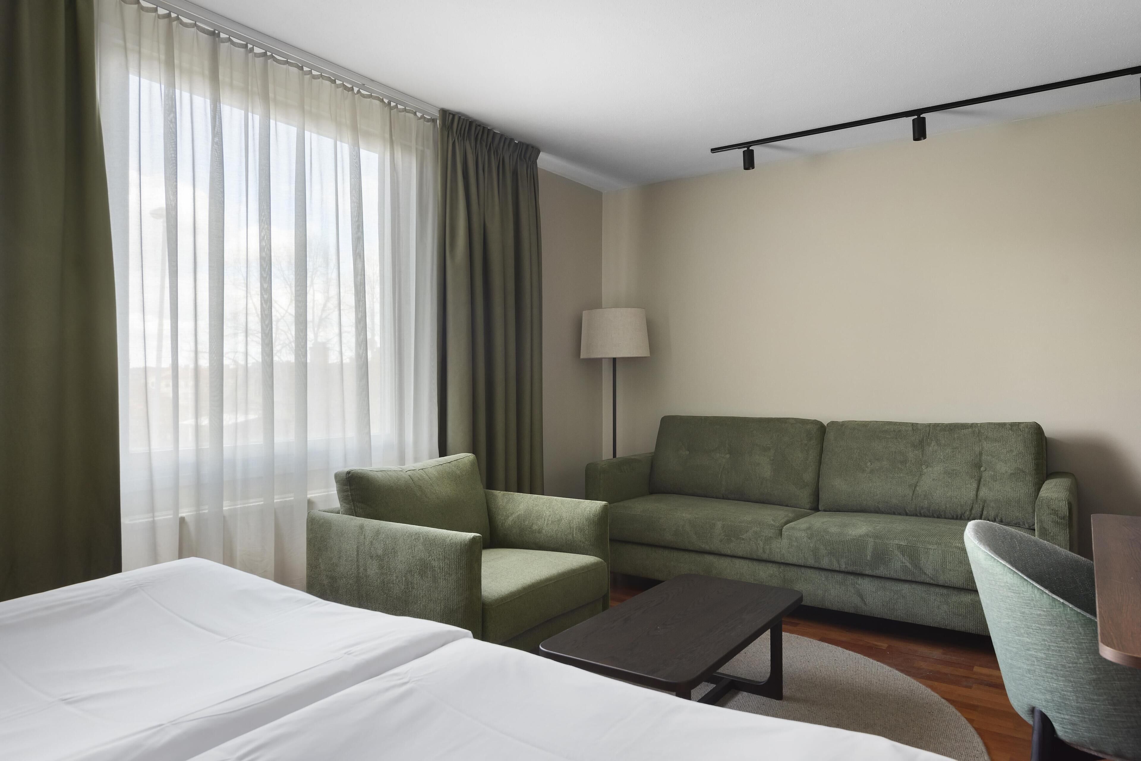Una habitación de hotel con una cama, un sofá verde, un sillón verde, una mesita y una lámpara.