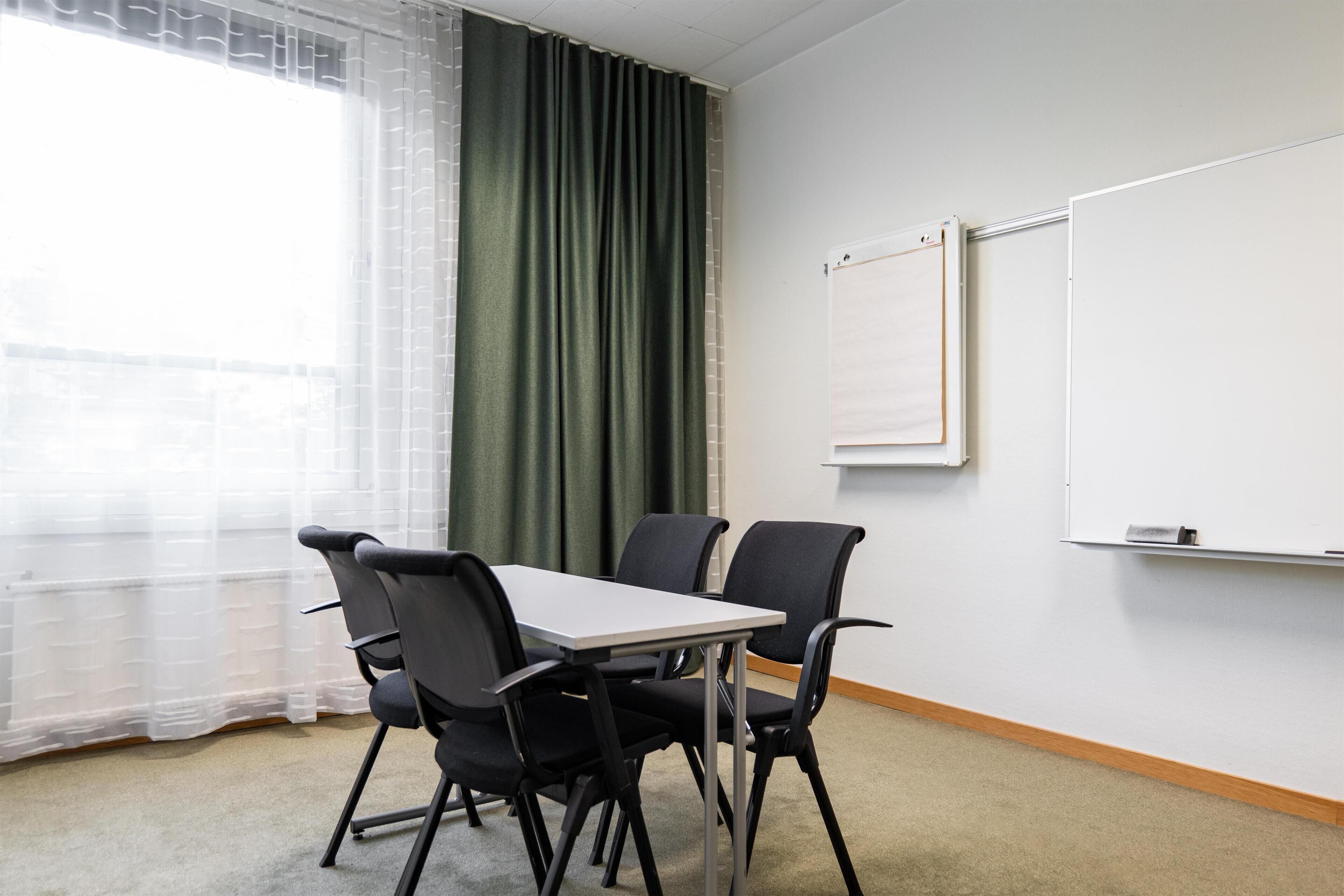 A conference room with a table and four chairs, a whiteboard, and a window with sheer curtains.