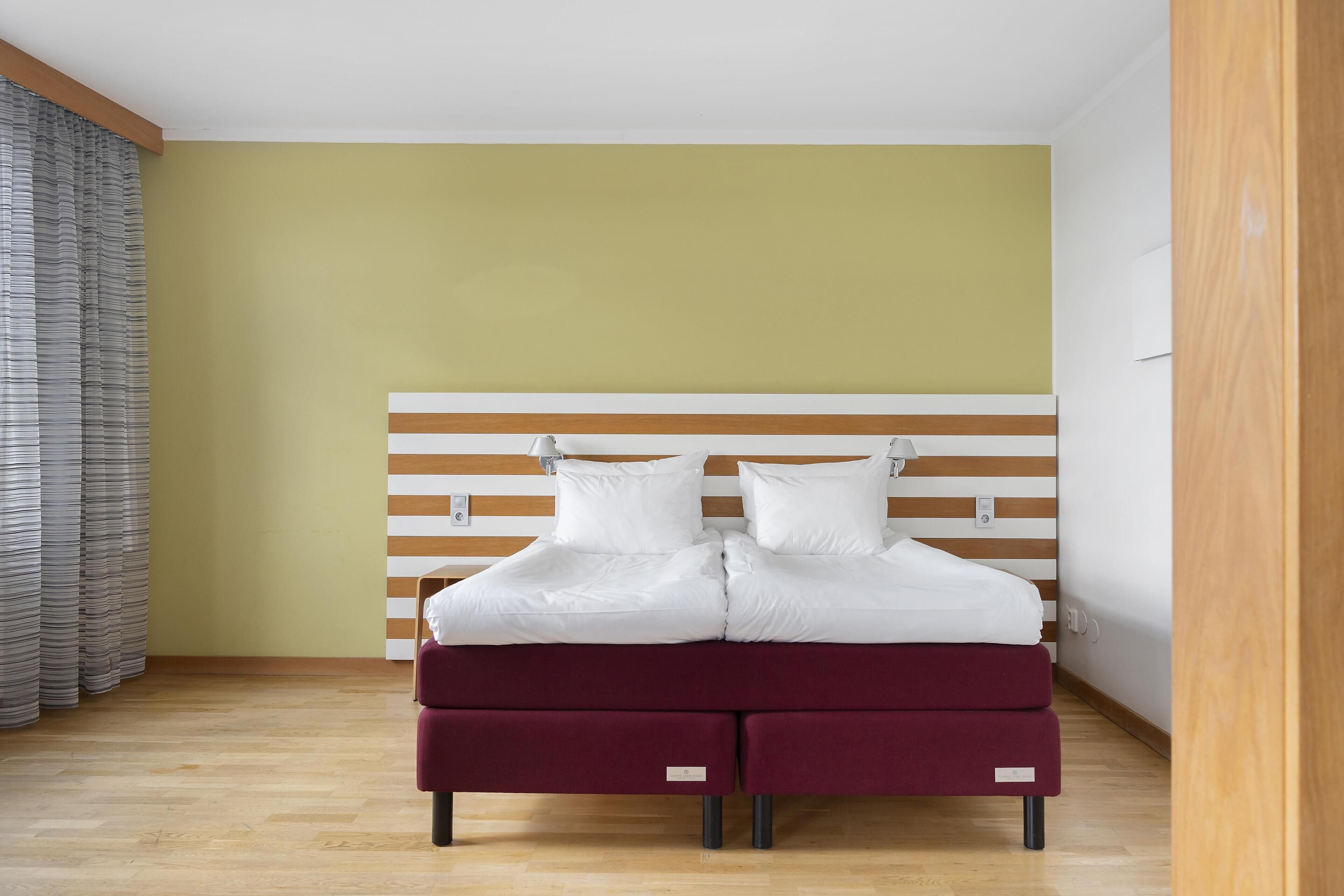 A double bed with a wooden headboard, a red footboard, and white bedding in a room with wooden flooring and a yellow wall.