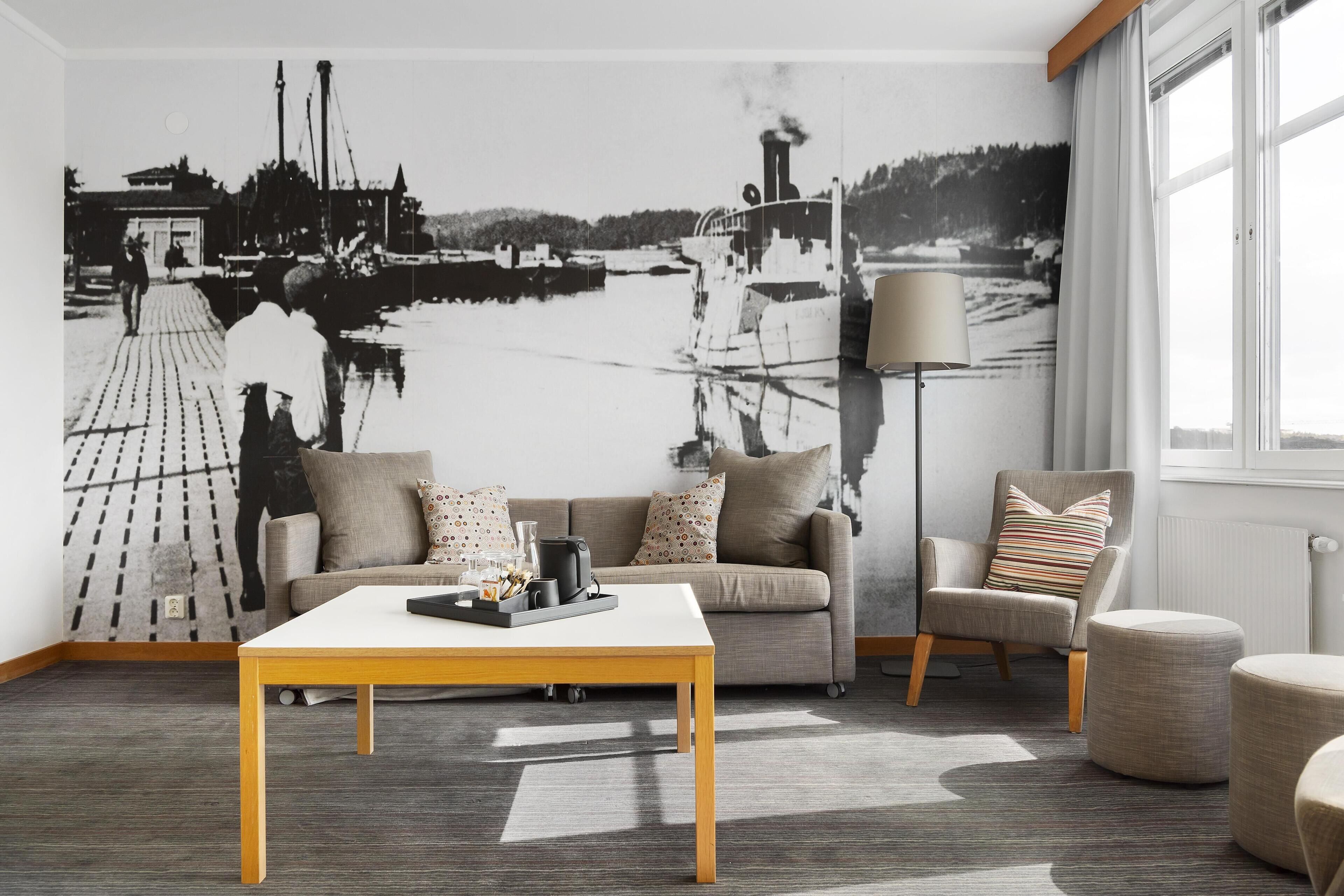 A living room with a sofa, armchair, coffee table, and a large wall mural of a harbor scene.