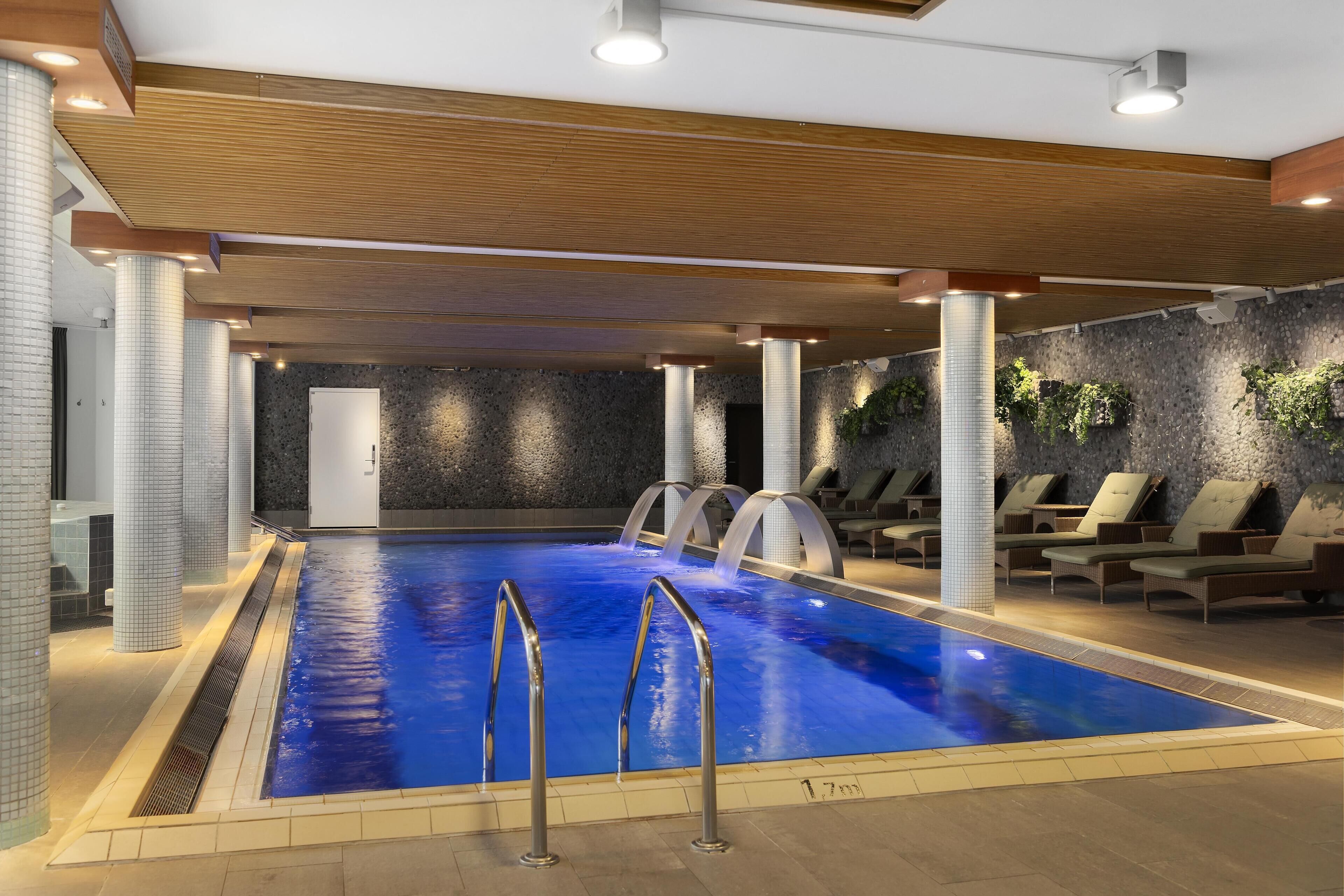 An indoor swimming pool with clear water, surrounded by lounge chairs and a stone wall.