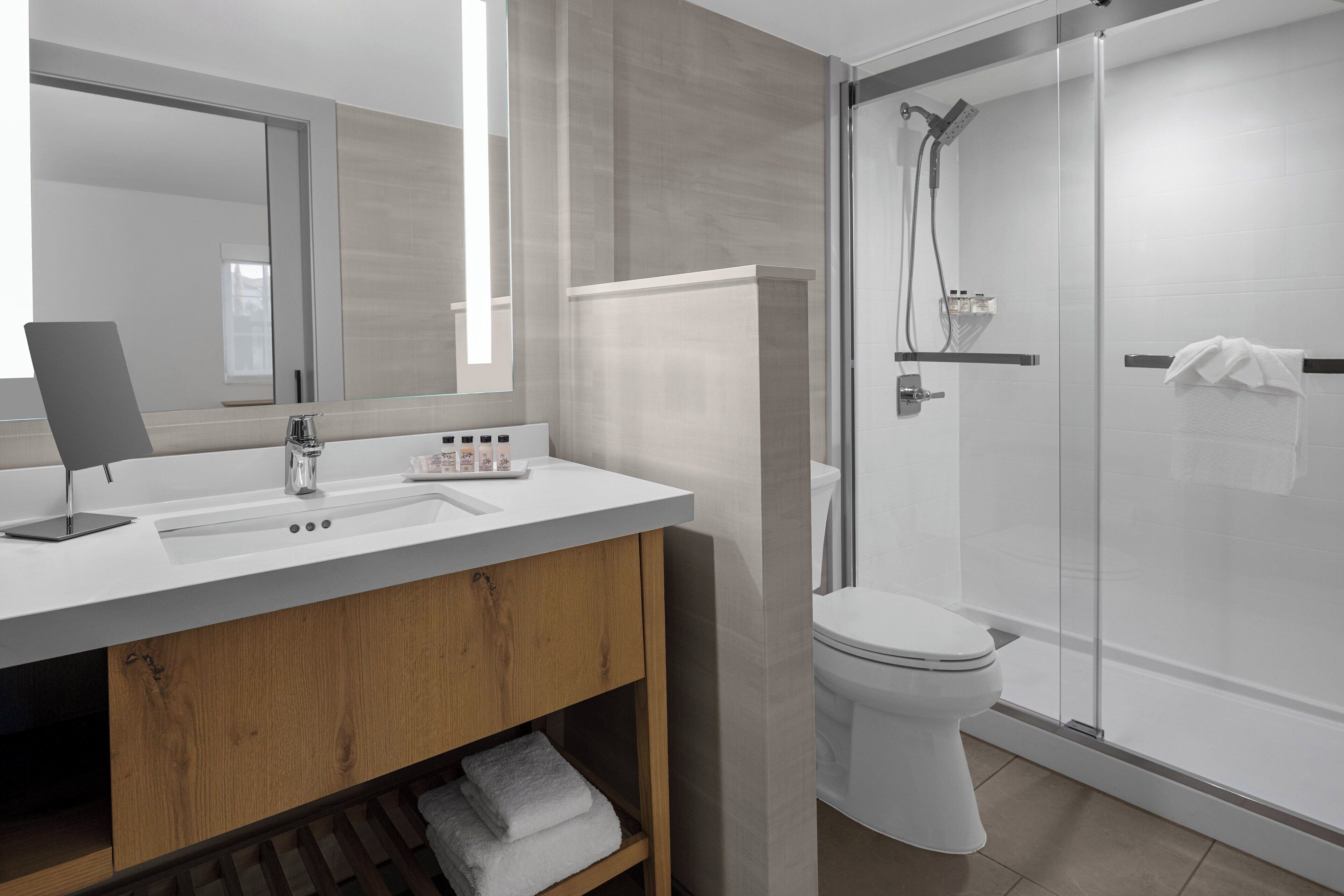 A modern bathroom with a white sink, mirror, and toilet. There is a glass shower enclosure and a wooden vanity with a towel rack.