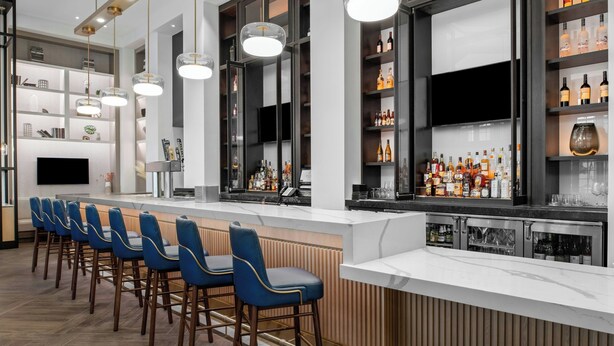 A modern bar area with a long counter, blue bar stools, and a variety of bottles and glasses on shelves. -  Delta Hotels by Marriott Santa Clara (Santa Clara)