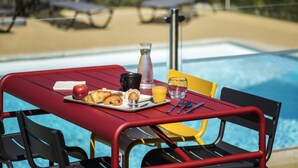 A dining table with breakfast items, a glass of orange juice, and a pitcher of water by a poolside. - ibis Roanne (Le Coteau)