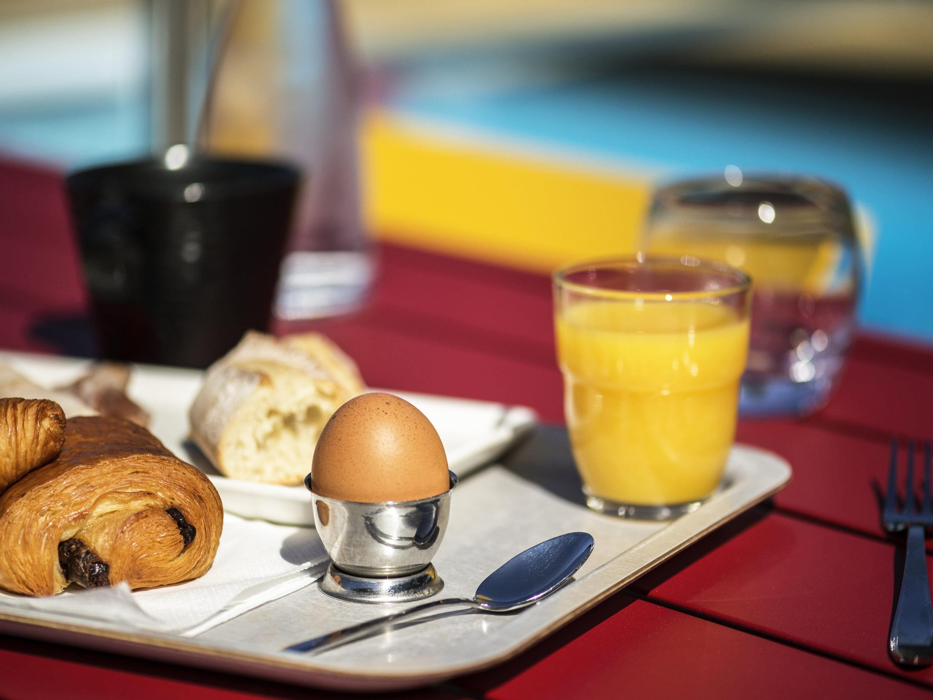 Een ontbijtserver met croissants, een ei, een glas sinaasappelsap en een vork op een rode tafelkleed.