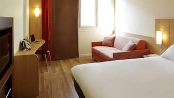 A hotel room with a bed, a desk with a laptop, a chair, a sofa, and a window with curtains.