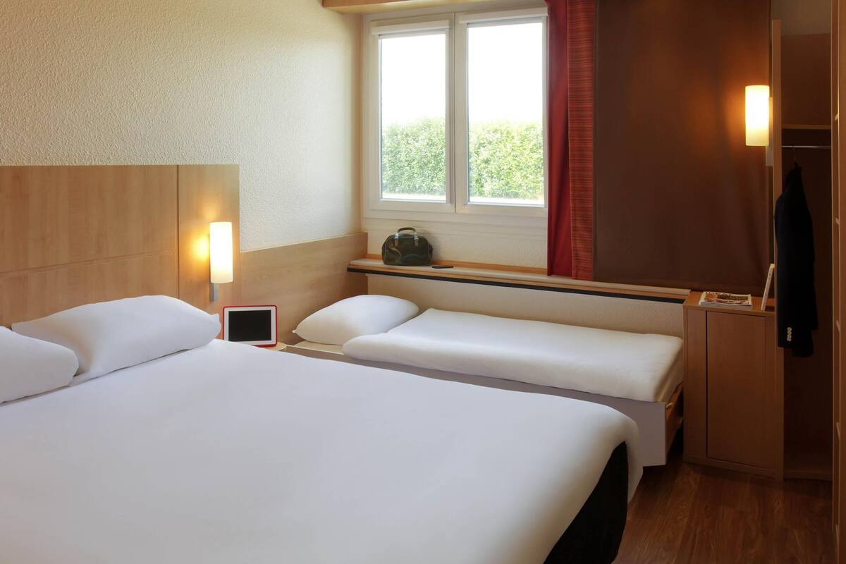 A hotel room with a large bed, bedside tables, a window with curtains, a coat rack, and a bedside lamp.