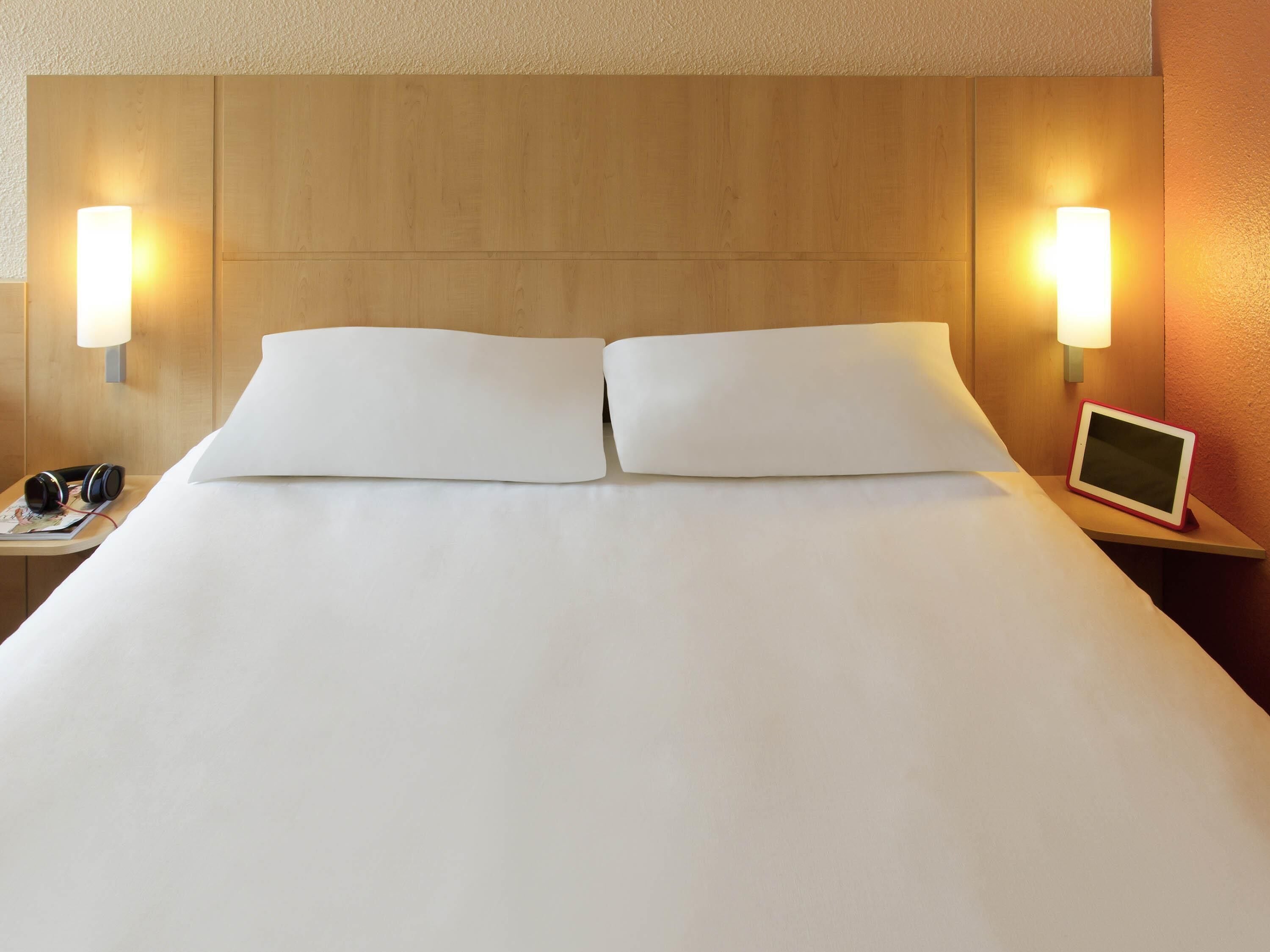 A hotel room with a large bed, two bedside lamps, a tablet on a nightstand, and a pair of headphones on the bed.