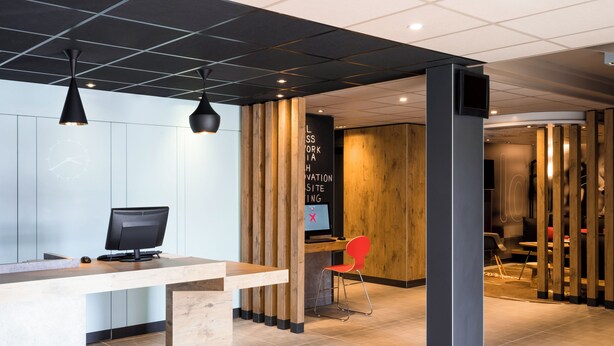A modern reception area with a wooden desk, a computer, and a red chair. - ibis Roanne (Le Coteau)