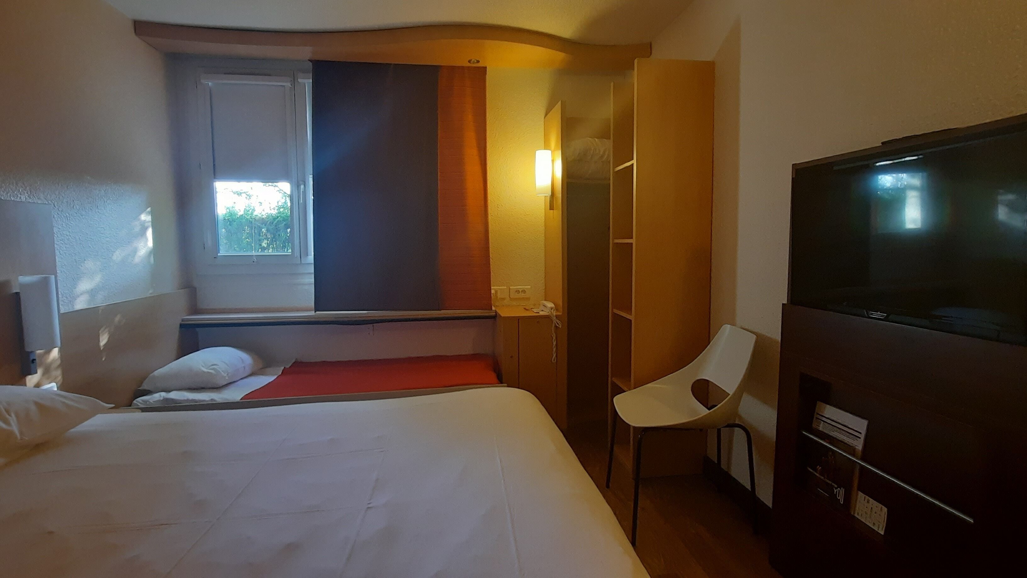 A hotel room with a bed, a chair, a TV, a window, and a wardrobe.