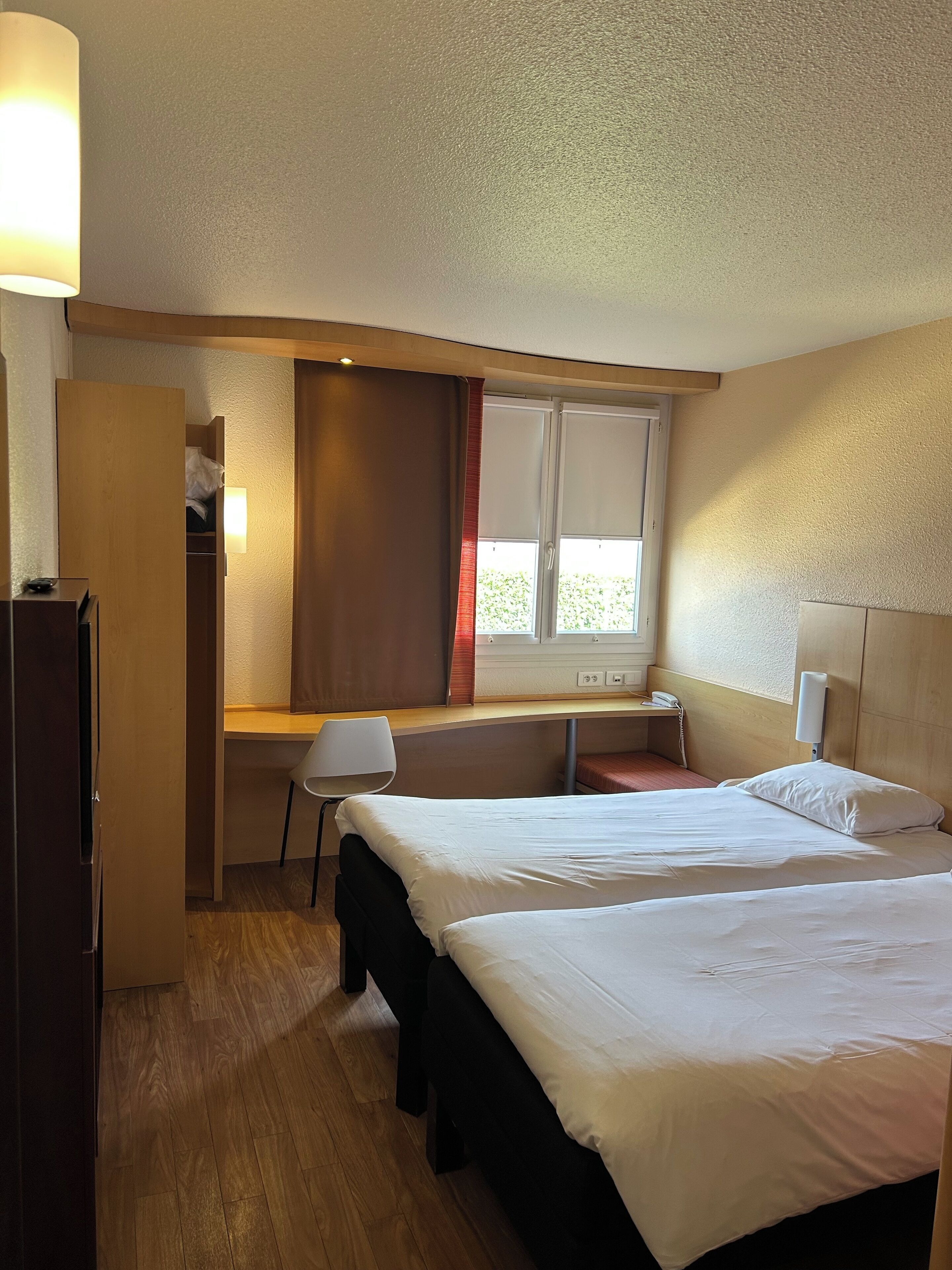 Une chambre d’hôtel comprenant un lit, un bureau, une chaise, une armoire et une fenêtre avec des rideaux.