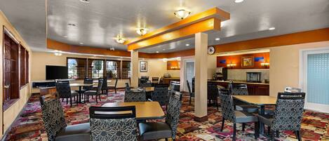 A hotel lobby with multiple dining tables and chairs, a kitchen area, and a reception desk.
