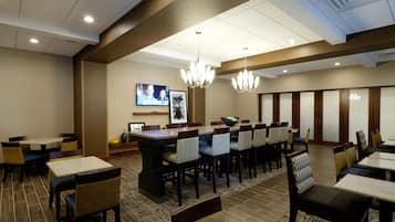 A hotel dining area with multiple tables and chairs, a television mounted on the wall, and a chandelier.