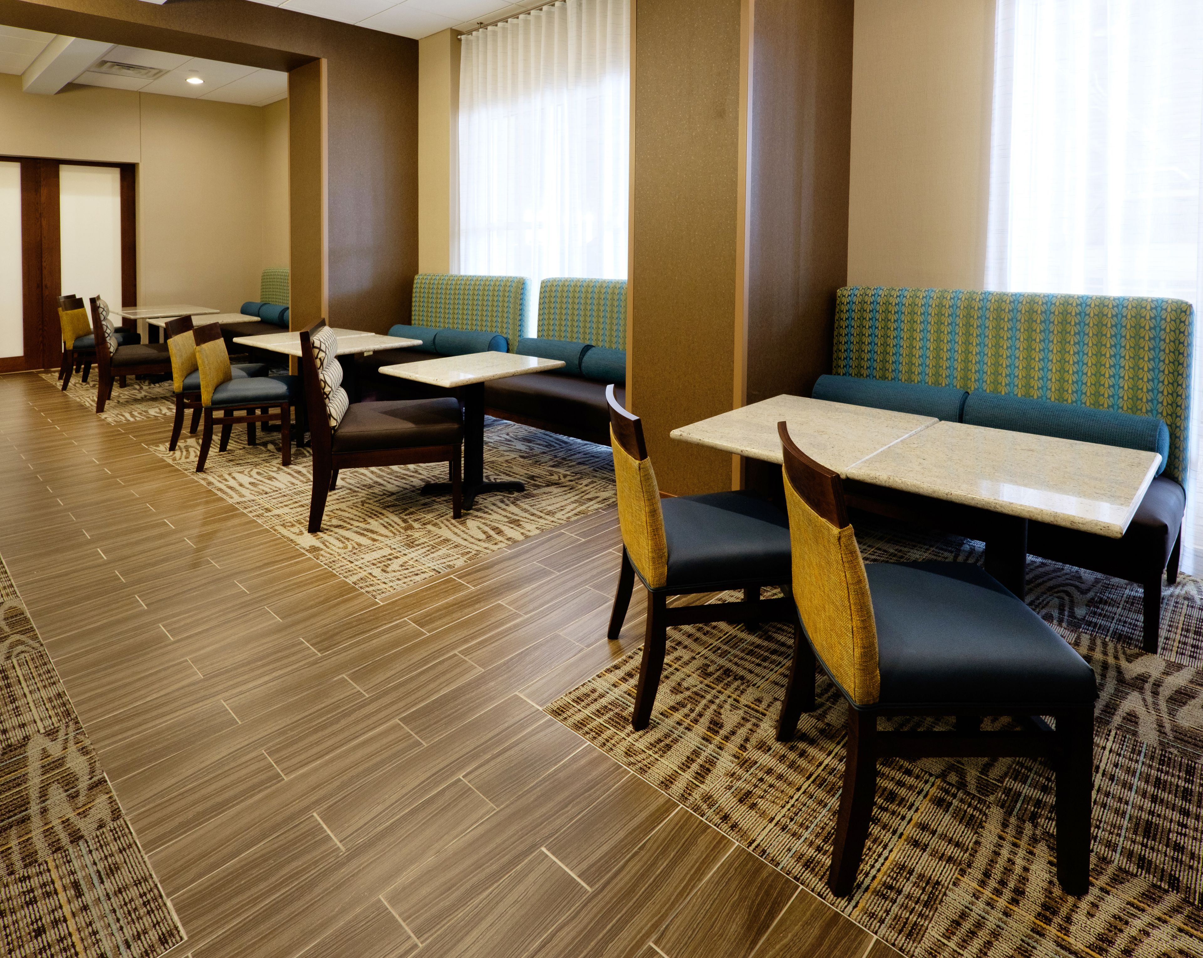 A hotel dining area with multiple tables and chairs, a patterned carpet, and large windows with curtains.