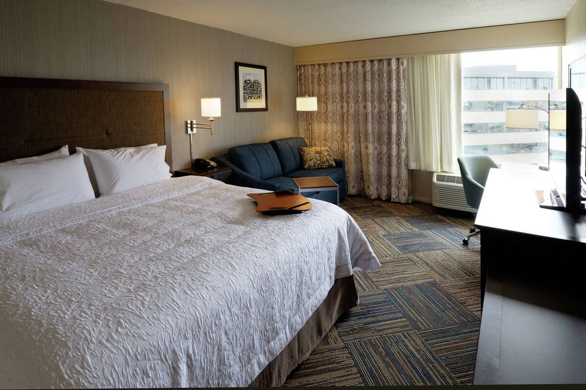A hotel room with a large bed, a desk, a chair, a sofa, and a window with curtains.