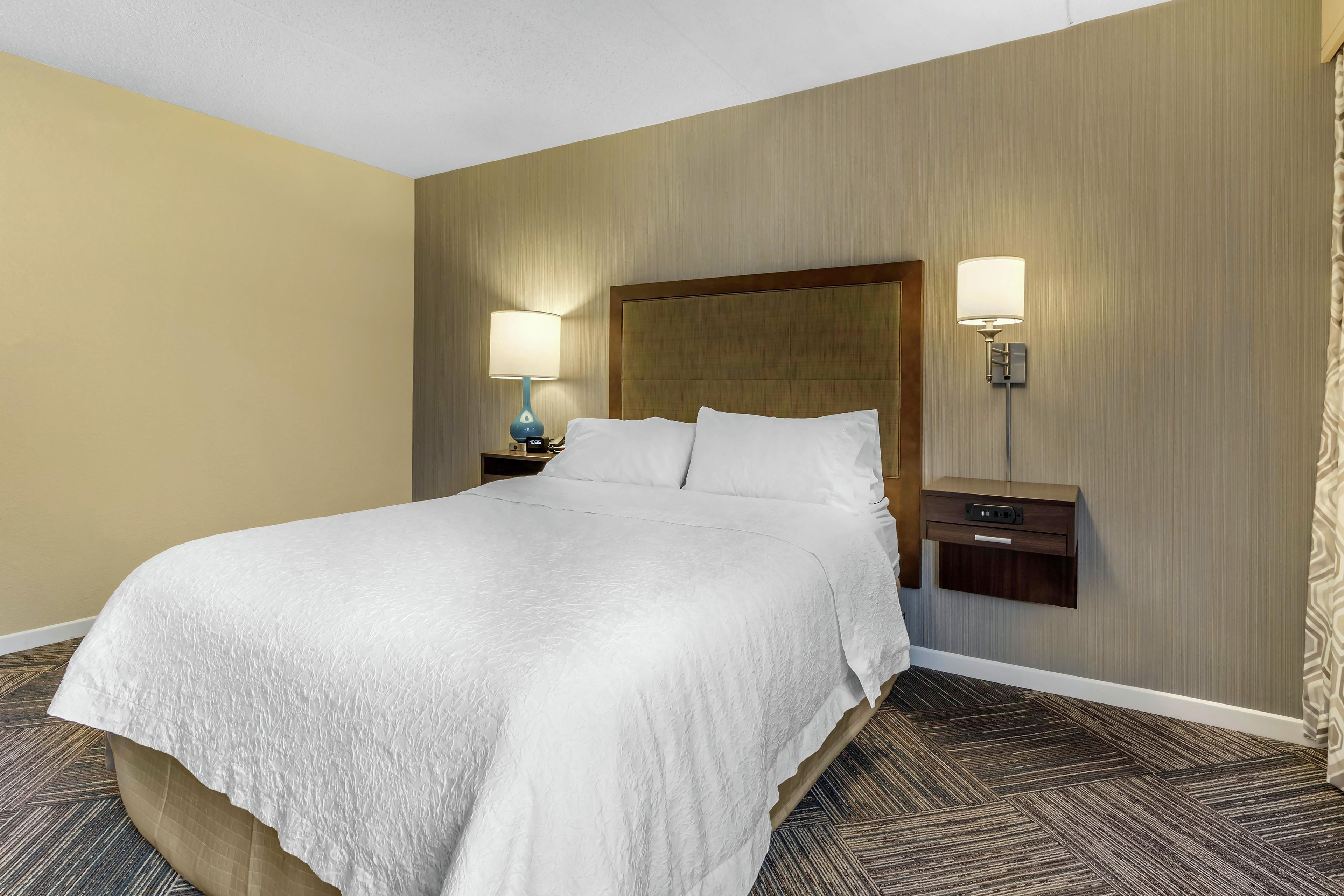 A hotel room with a large bed, two bedside tables, a nightstand, and a wall-mounted lamp.