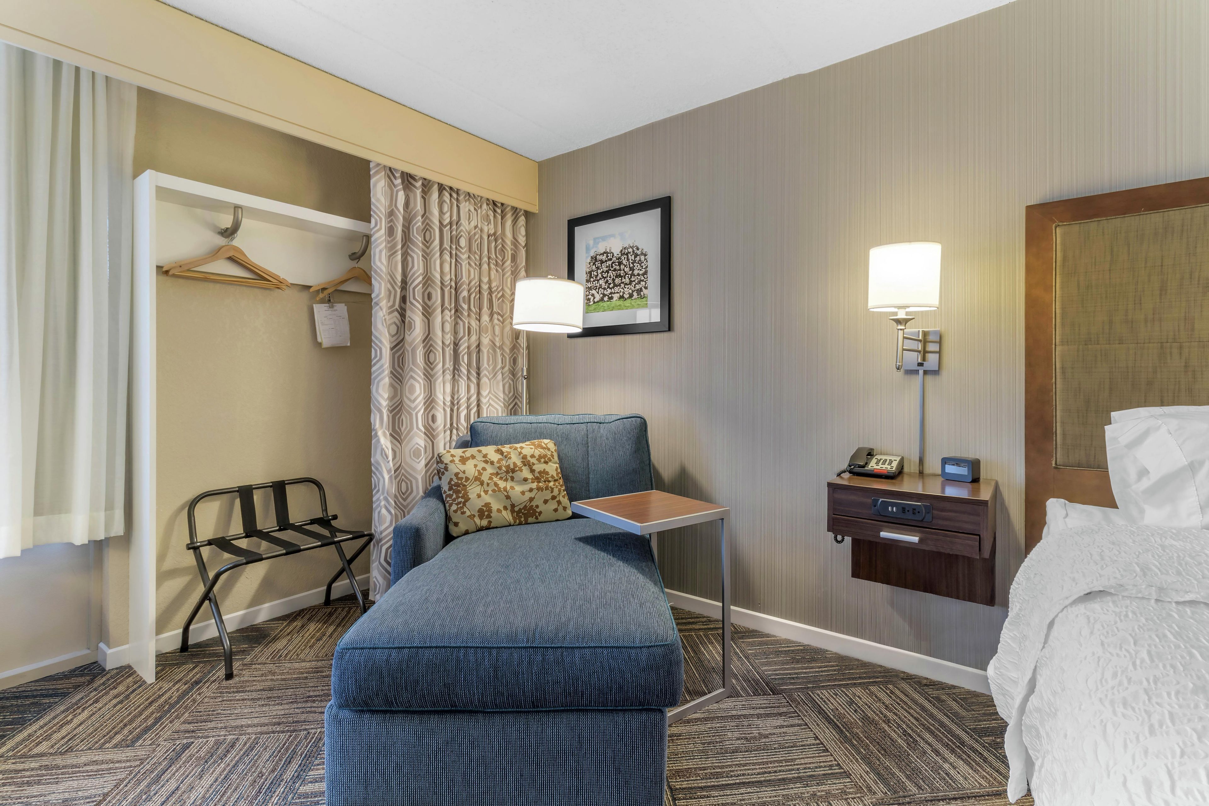 A hotel room with a bed, a desk with a lamp, a chair, a hanging coat rack, and a patterned curtain.