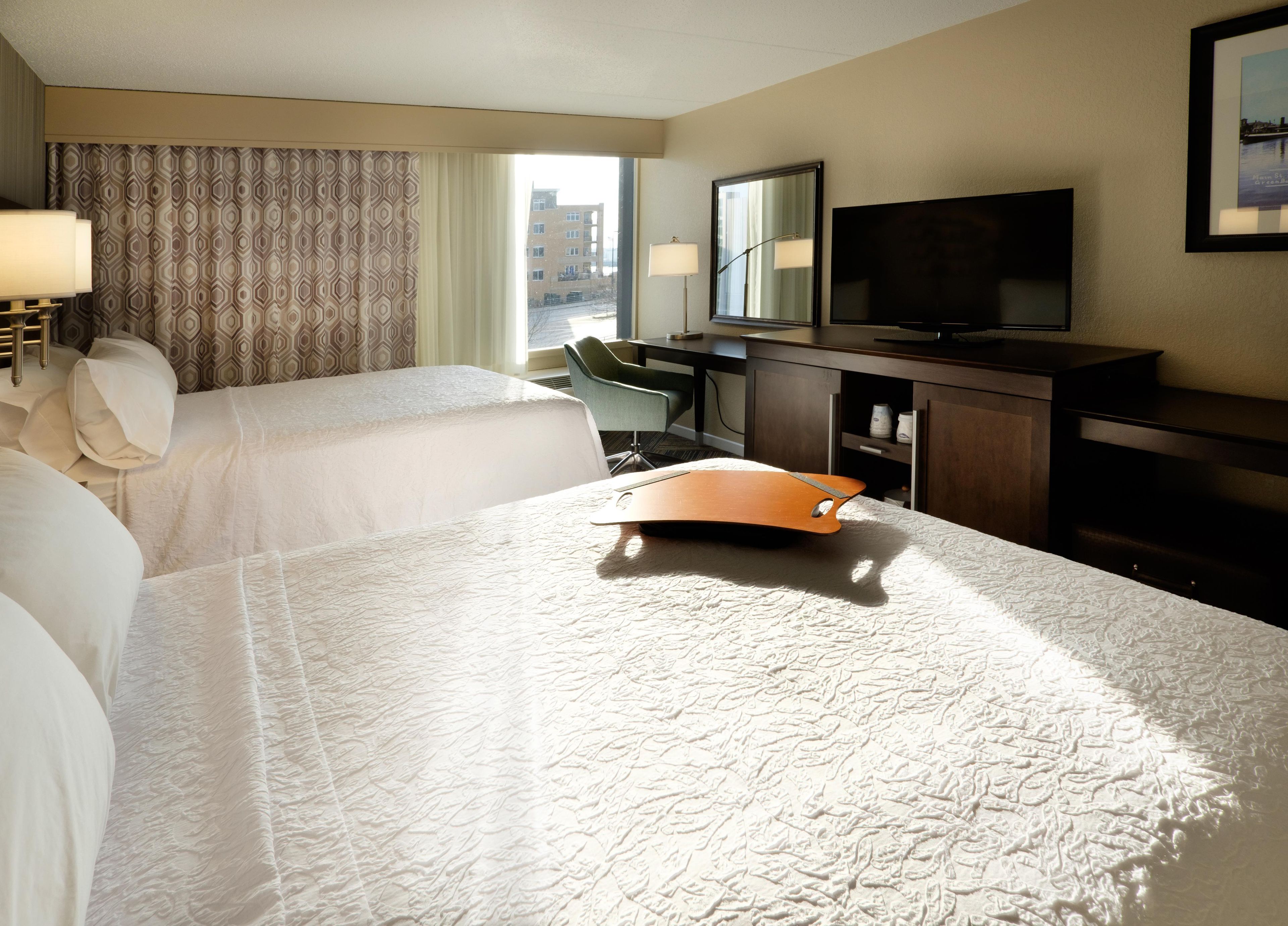 A hotel room with two beds, a desk, a chair, a television, and a window with a view of buildings.