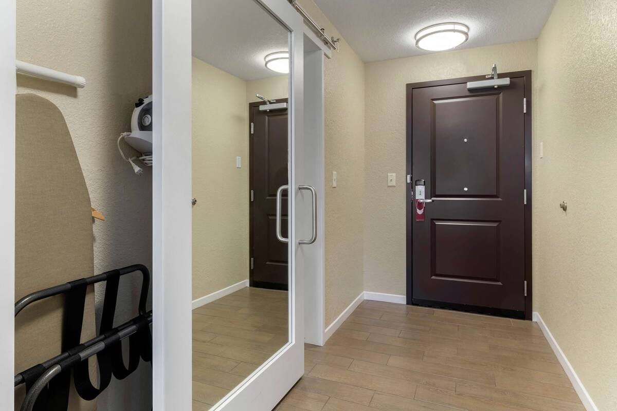 A hotel hallway with a mirrored wall, a door with a handle, and a small seating area with a chair and a bench.