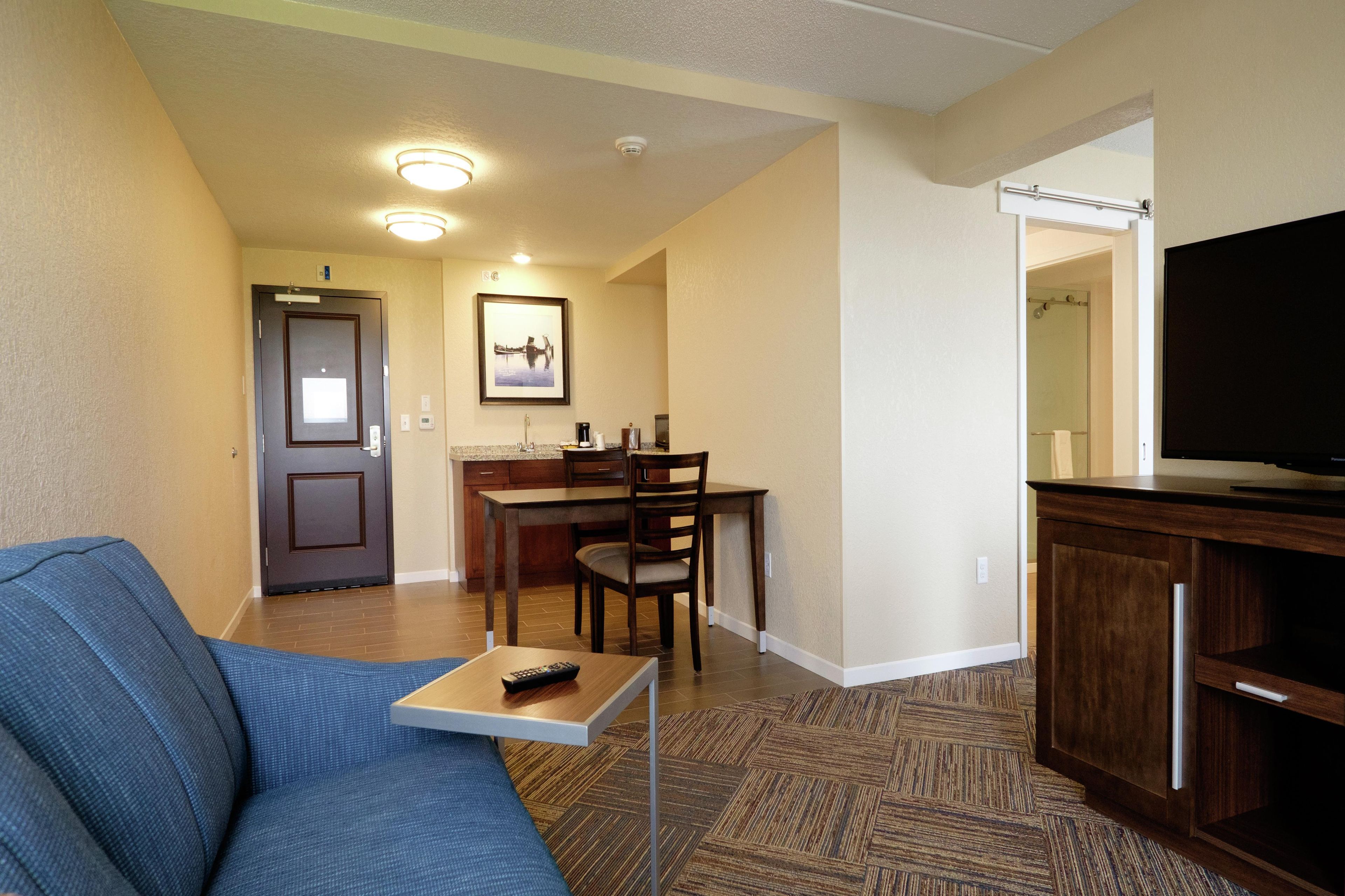Een hotelkamer met een bank, een klein tafeltje met een afstandsbediening, een zithoek met een stoel en een tafel, een televisie op een dressoir en een deur naar een andere kamer.