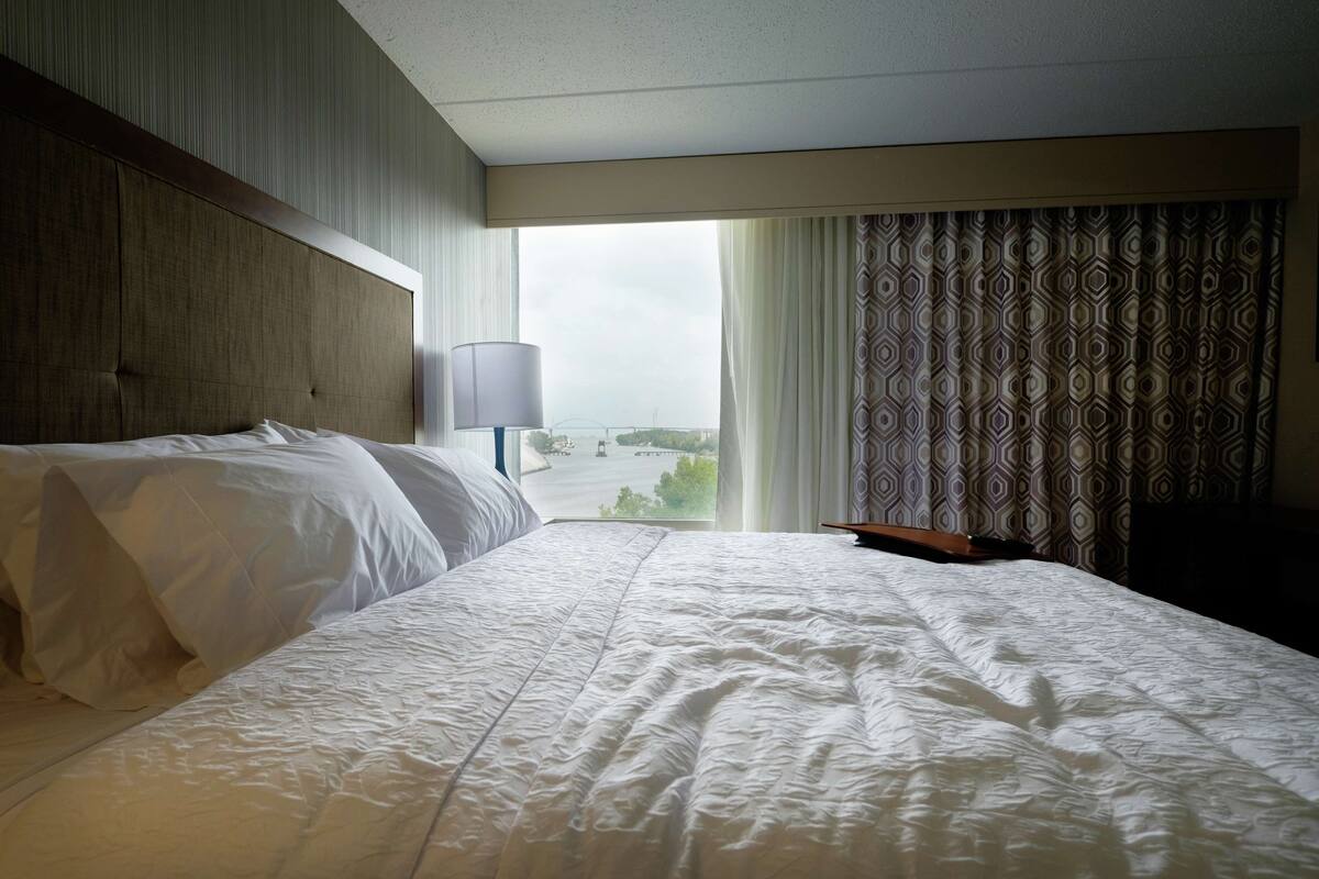 A hotel room with a large bed, a headboard, a window with curtains, and a view of the outside.