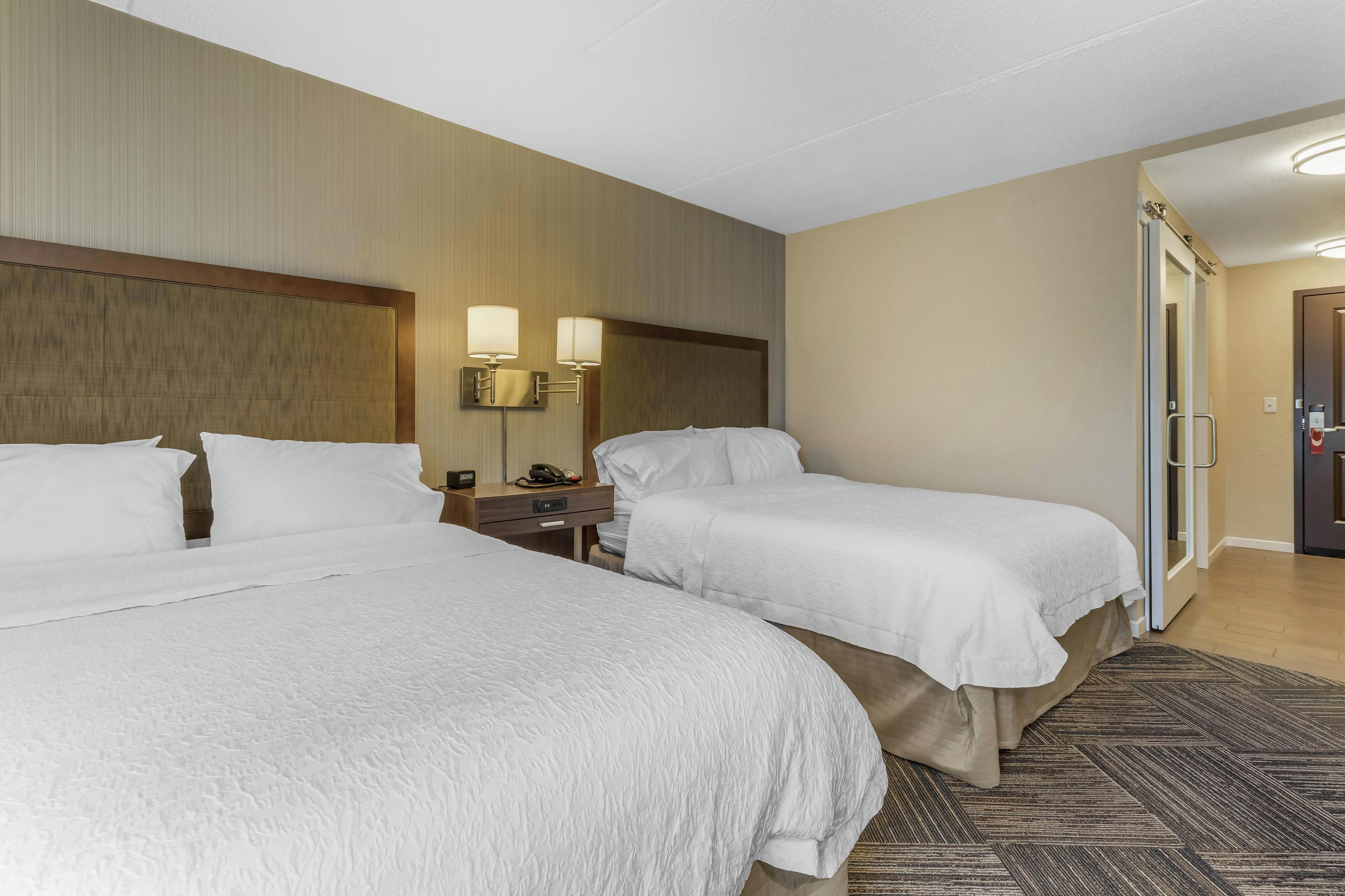 A hotel room with two beds, a nightstand with a phone and a lamp, and a door leading to a bathroom.