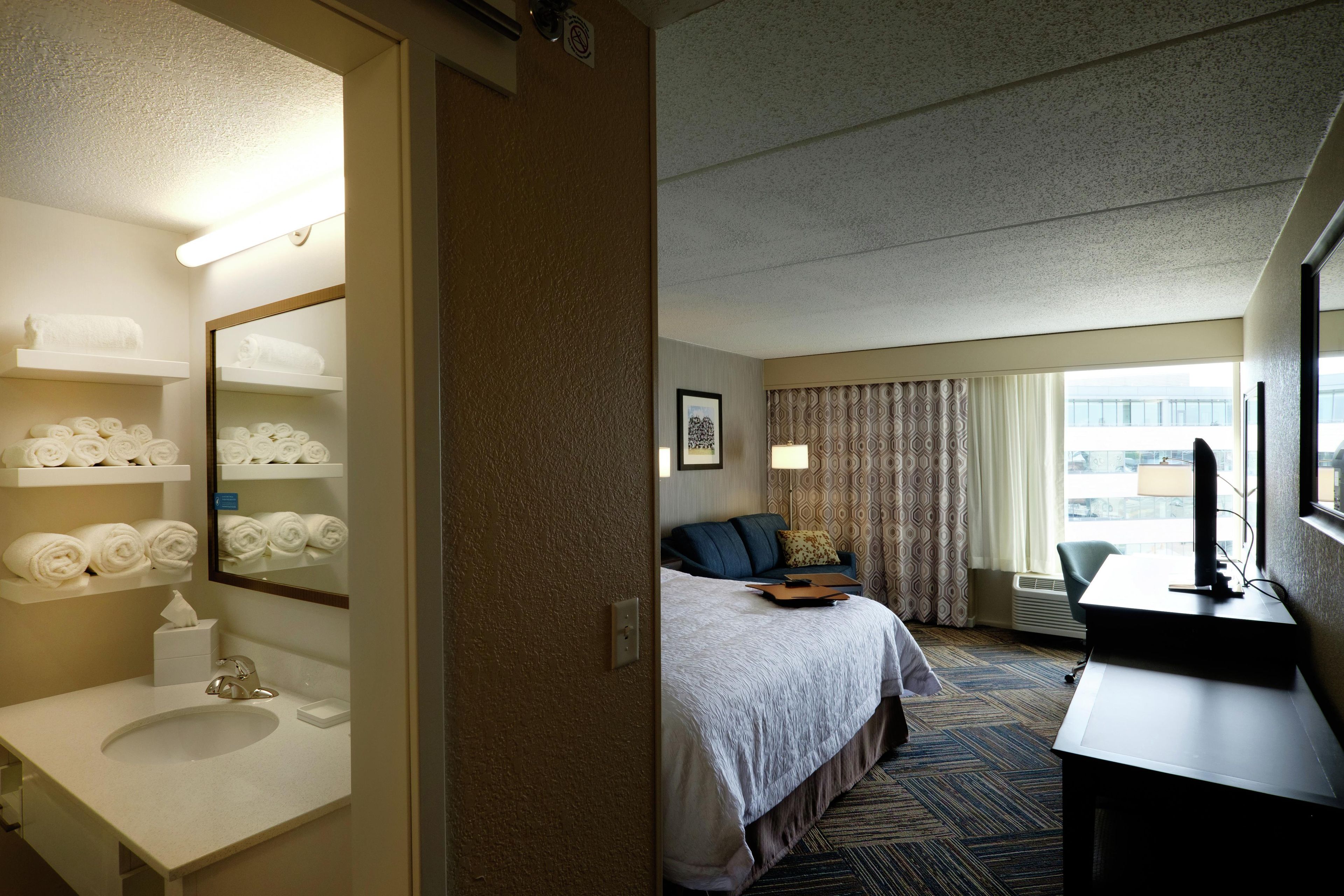A hotel room with a bed, a desk, a chair, a bathroom with a sink and towel rack.
