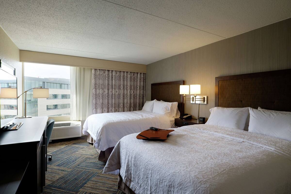 A hotel room with two beds, a desk, and a large window.