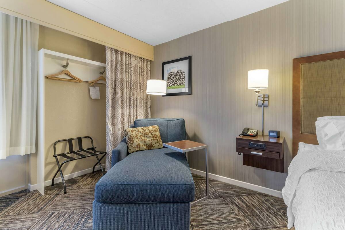A hotel room with a blue sofa, a small table, a bedside table with a lamp, a coat rack, and a bed with white bedding.
