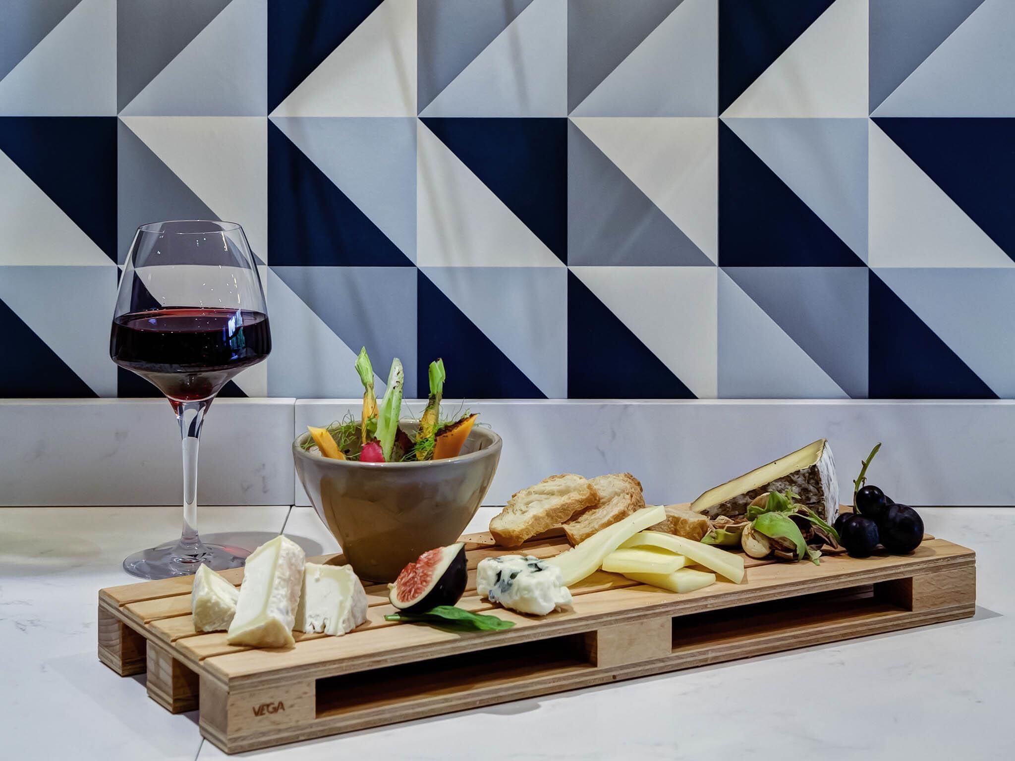 Une planche en bois sur laquelle sont disposés différents fromages, un saladier rempli de salade et un verre de vin rouge, le tout posé sur une table devant un mur orné d’un motif géométrique.