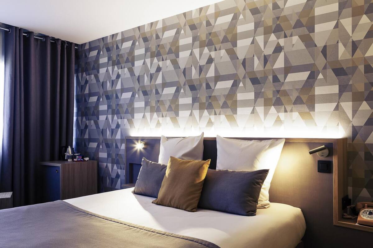 A modern hotel room with a geometric-patterned wall, a large bed with multiple pillows, a bedside table with a lamp, and a nightstand with a phone and a cup.