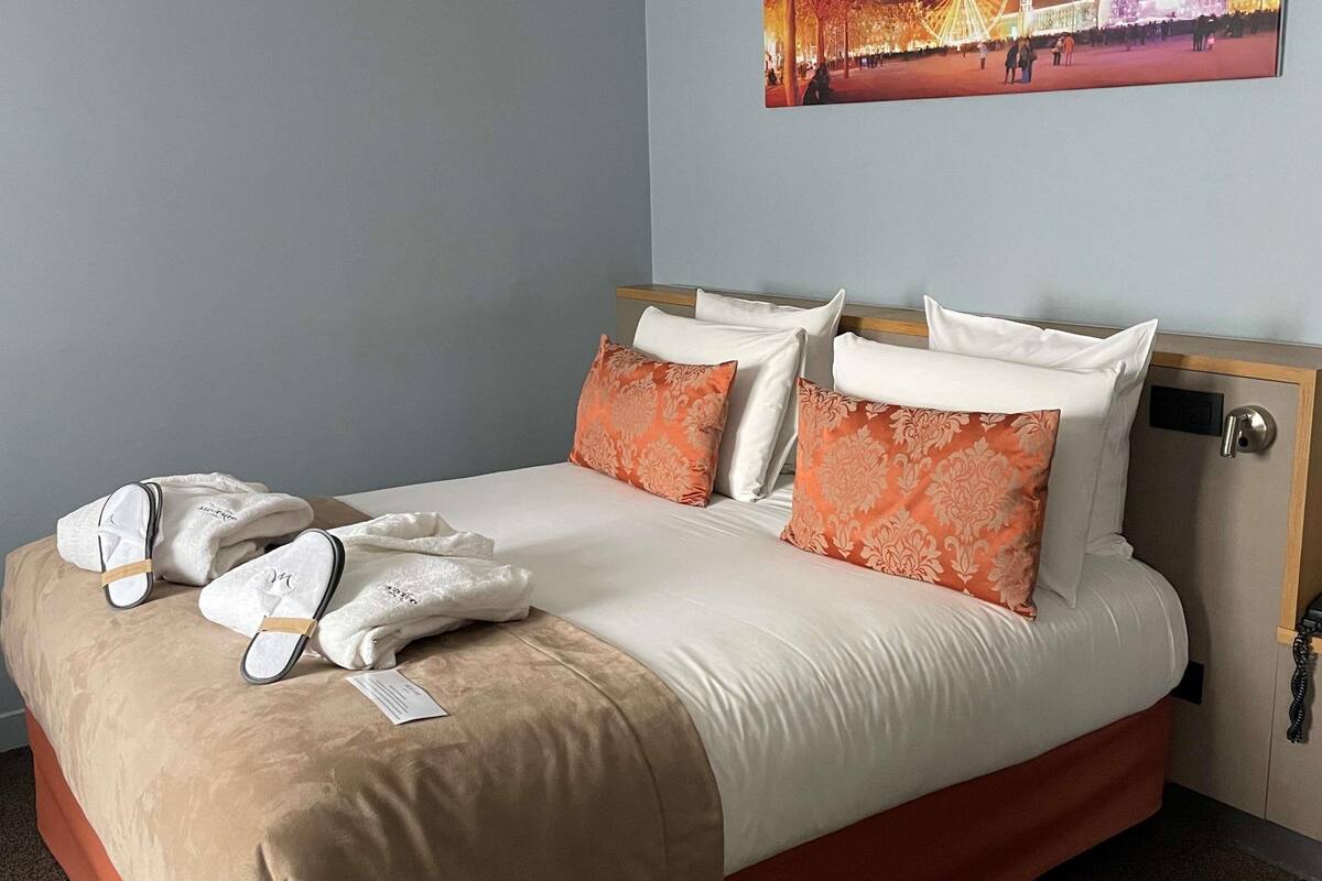 A hotel room with a bed, pillows, a blanket, and a coat rack with a jacket and a bag.