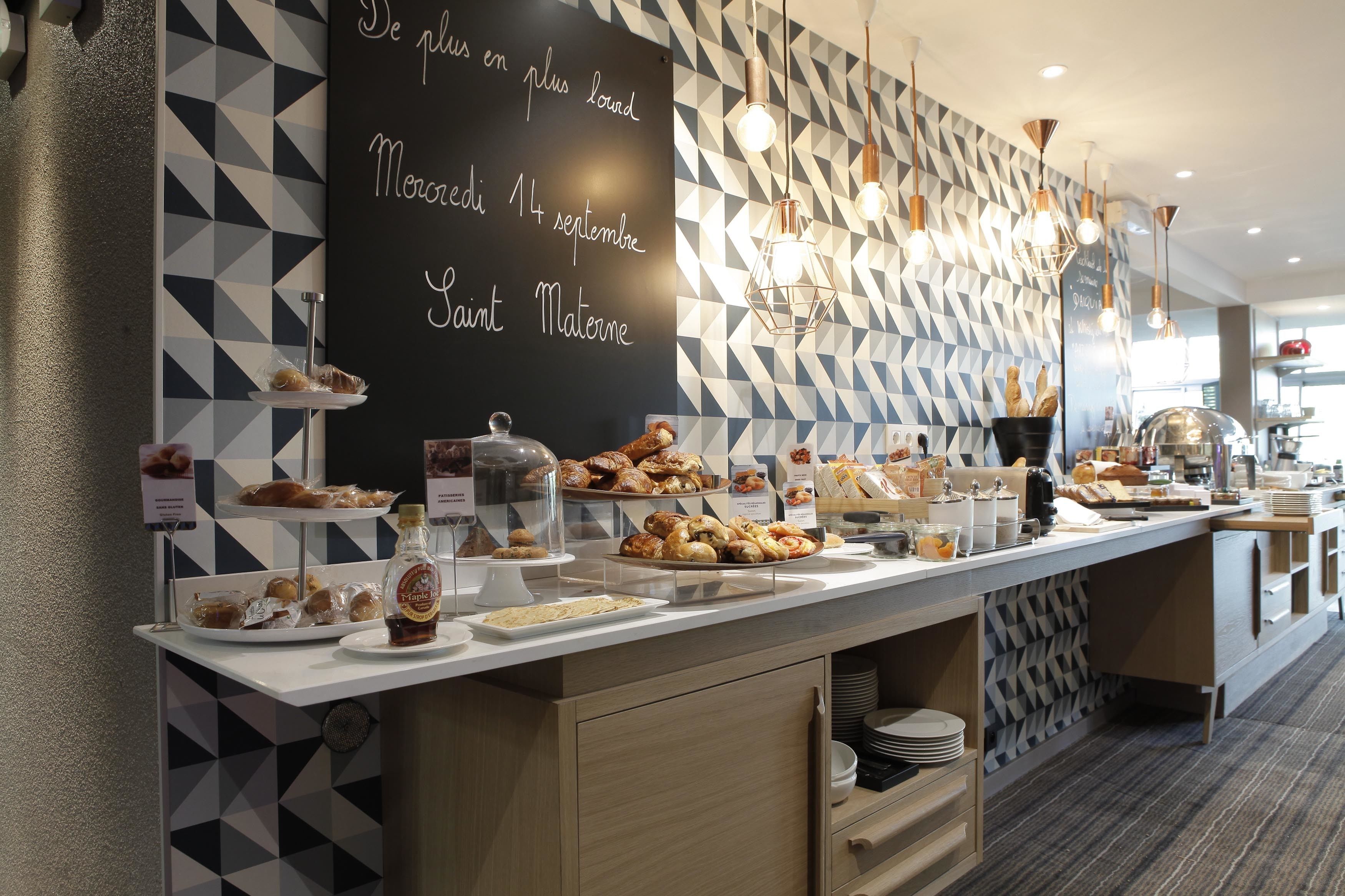 A breakfast buffet with various pastries and bread, a chalkboard with a message, and geometric patterned walls.