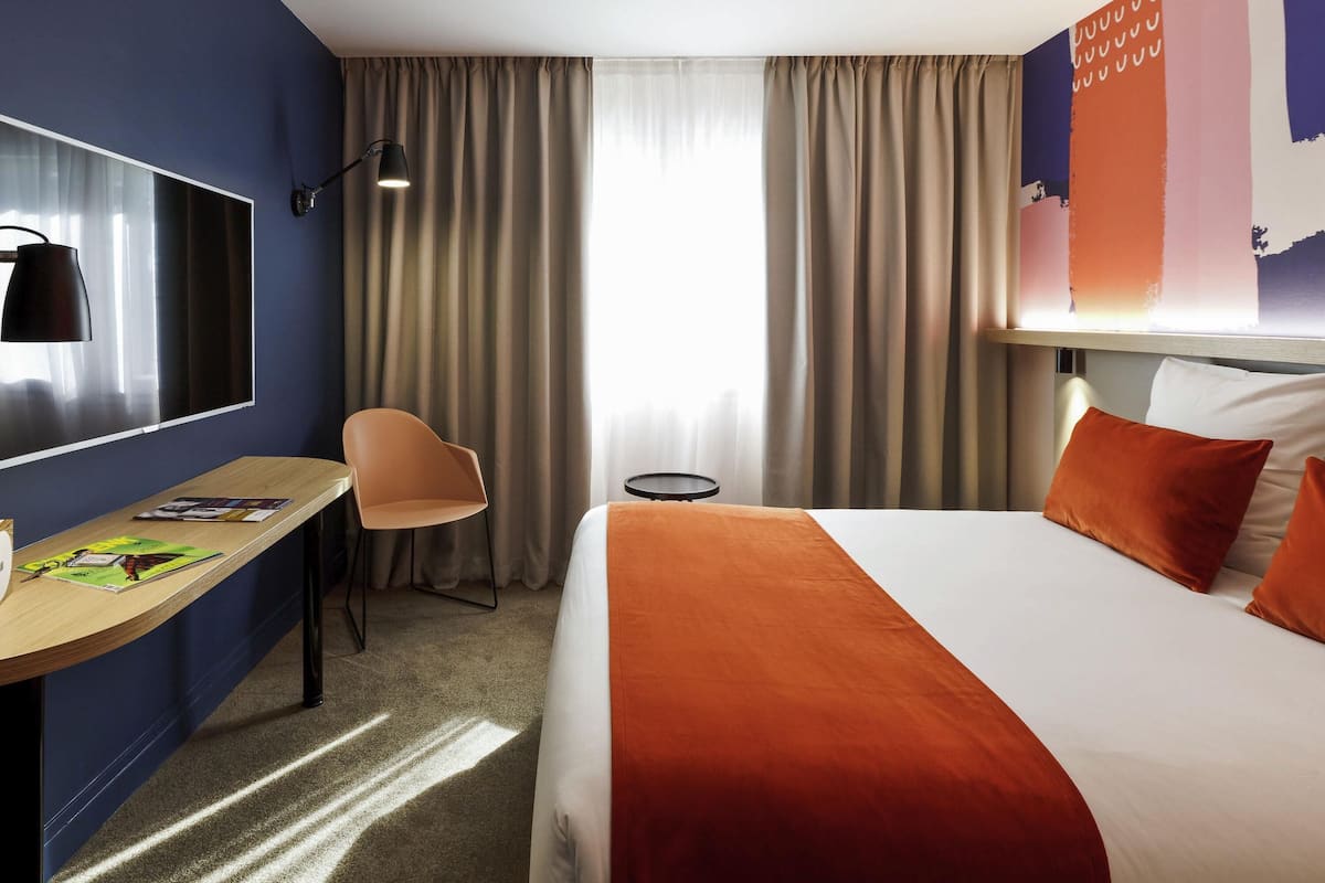 Une chambre d’hôtel avec un lit, un bureau, une chaise, une télévision et des rideaux.
