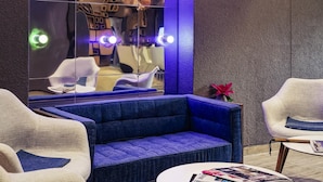 Un área de descanso moderna con un sofá azul, sillones blancos, una mesa redonda y un espejo grande que refleja la habitación.