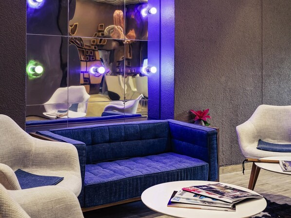Uma área de estar moderna com um sofá azul, poltronas brancas, uma mesa de centro redonda e um espelho grande refletindo o espaço.