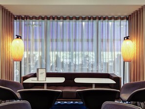 A hotel room with a dining area featuring a table and chairs, large windows with curtains, and two wall-mounted lamps. - Mercure Lyon Charbonnieres (Charbonnieres-les-Bains)