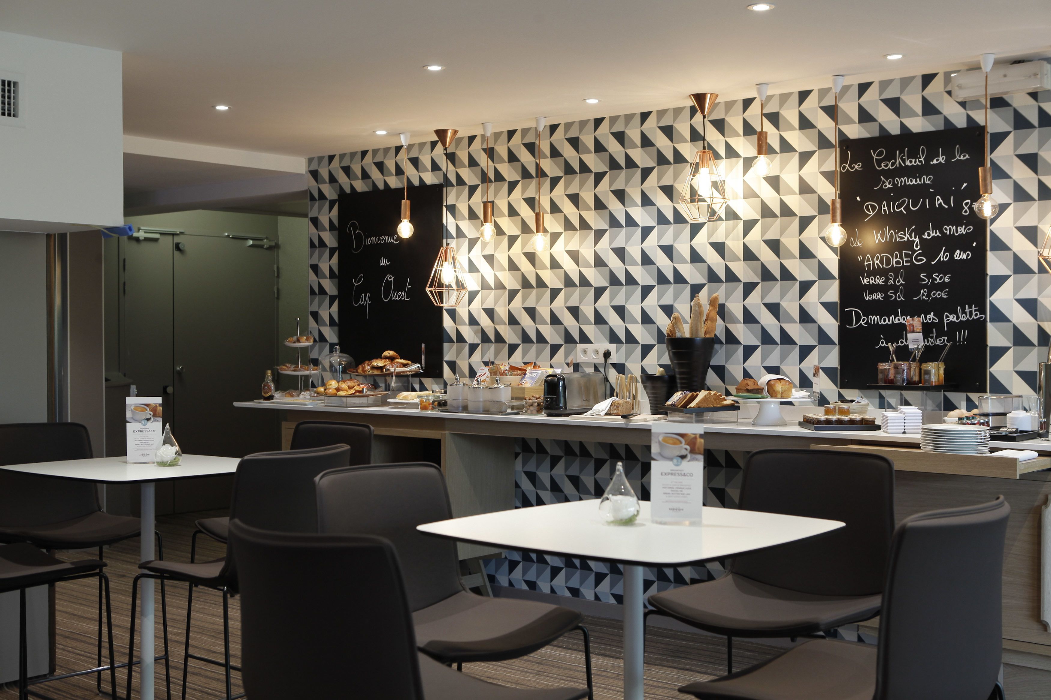 A modern cafe with a geometric patterned wall, a bar area with food and drinks, and several tables and chairs.