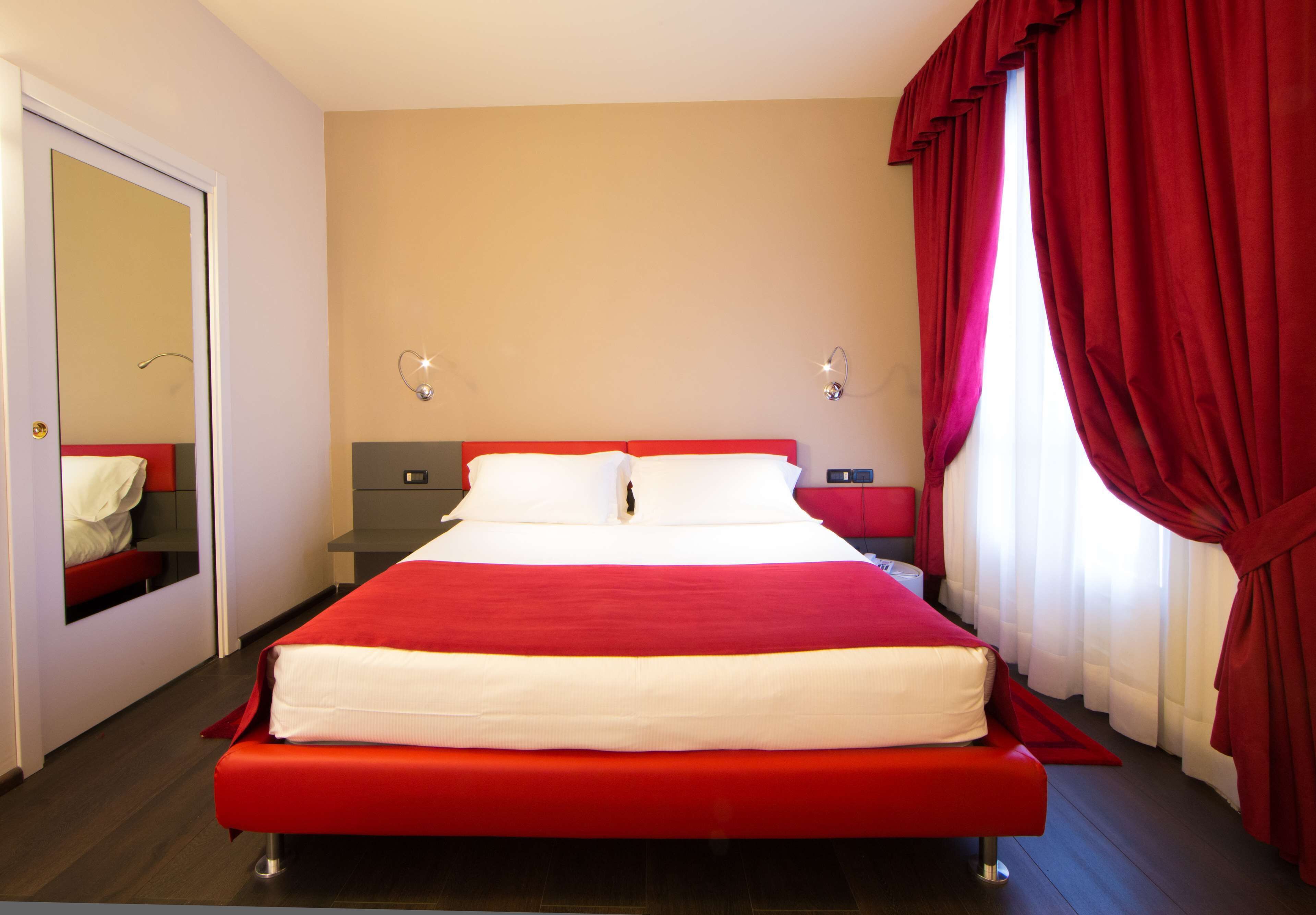 Une chambre d’hôtel avec un grand lit, une table de chevet, un miroir et des rideaux rouges.