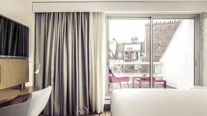 A hotel room with a bed, a desk, a chair, a TV, and a balcony with a view of buildings. - Mercure Paris Arc de Triomphe Etoile (Paris)