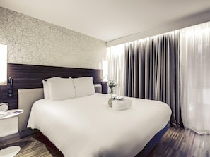Quarto de hotel com uma cama grande, uma mesa de cabeceira com abajur, telefone na parede e janela com cortinas.