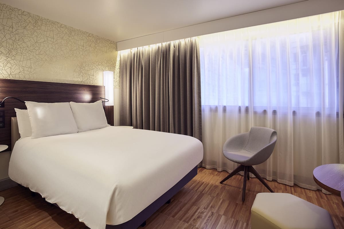 Ein Hotelzimmer mit einem großen Bett, einem Sessel, einem kleinen Tisch und einem Fenster mit Vorhängen.
