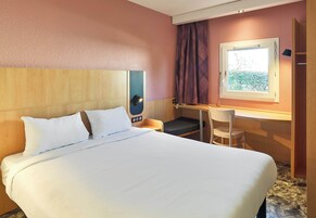 Habitación de hotel con una cama grande, un escritorio con silla, una ventana con cortinas y un cabecero con panel de control.