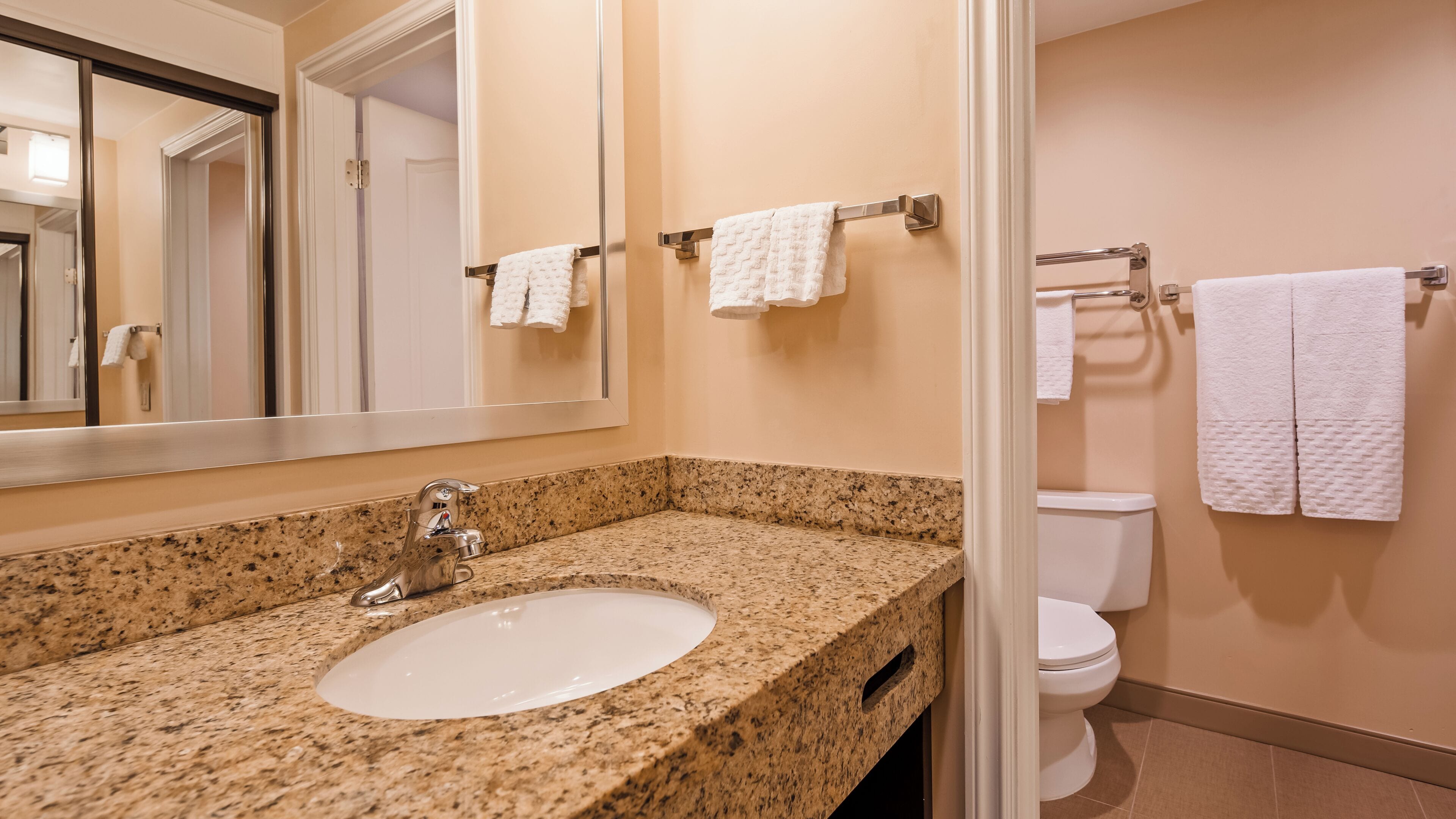 Baño con lavamanos de granito, espejo, inodoro y dos toalleros con toallas blancas.