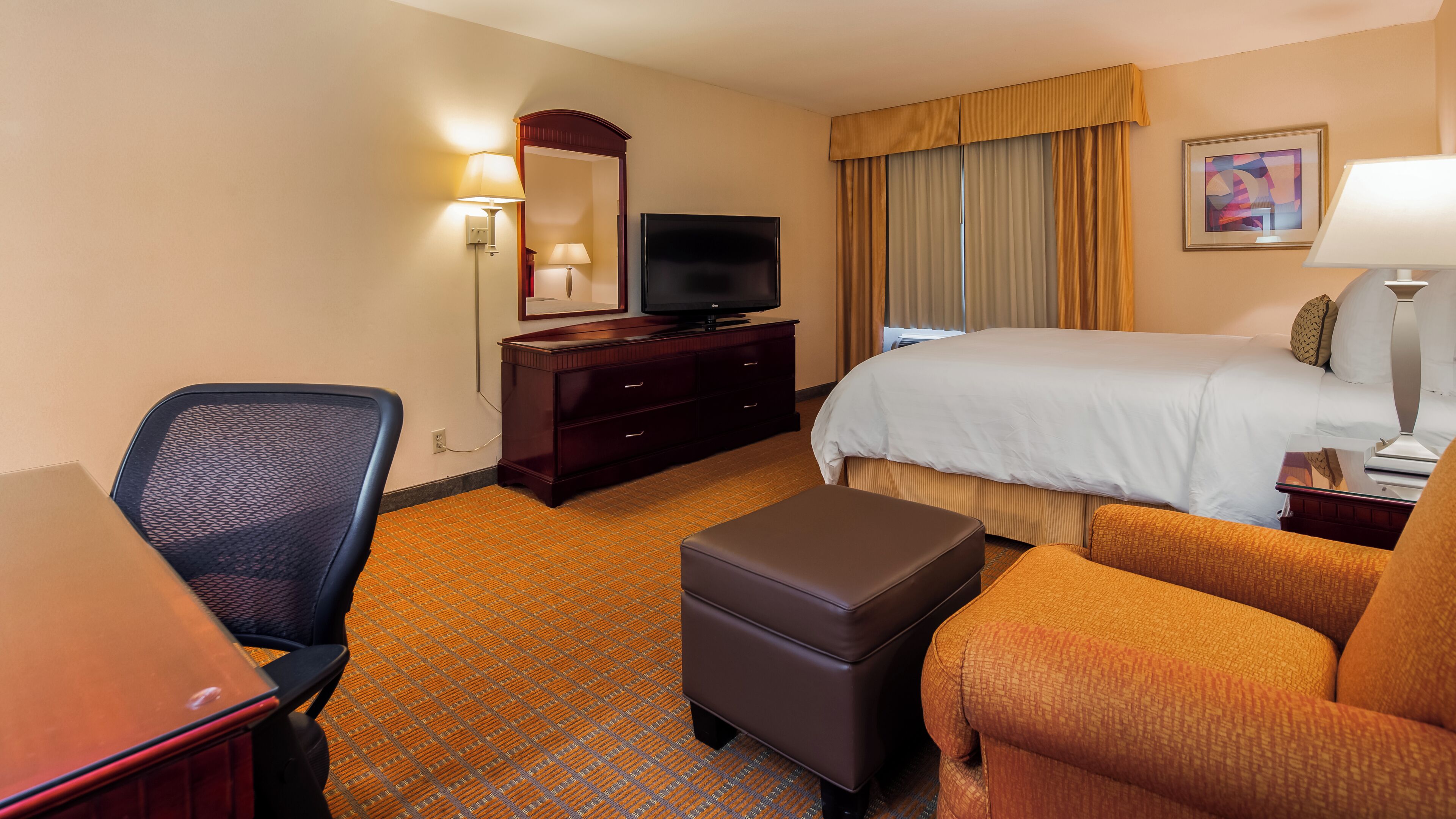 Quarto de hotel com cama, escrivaninha, cadeira, televisão e cômoda.